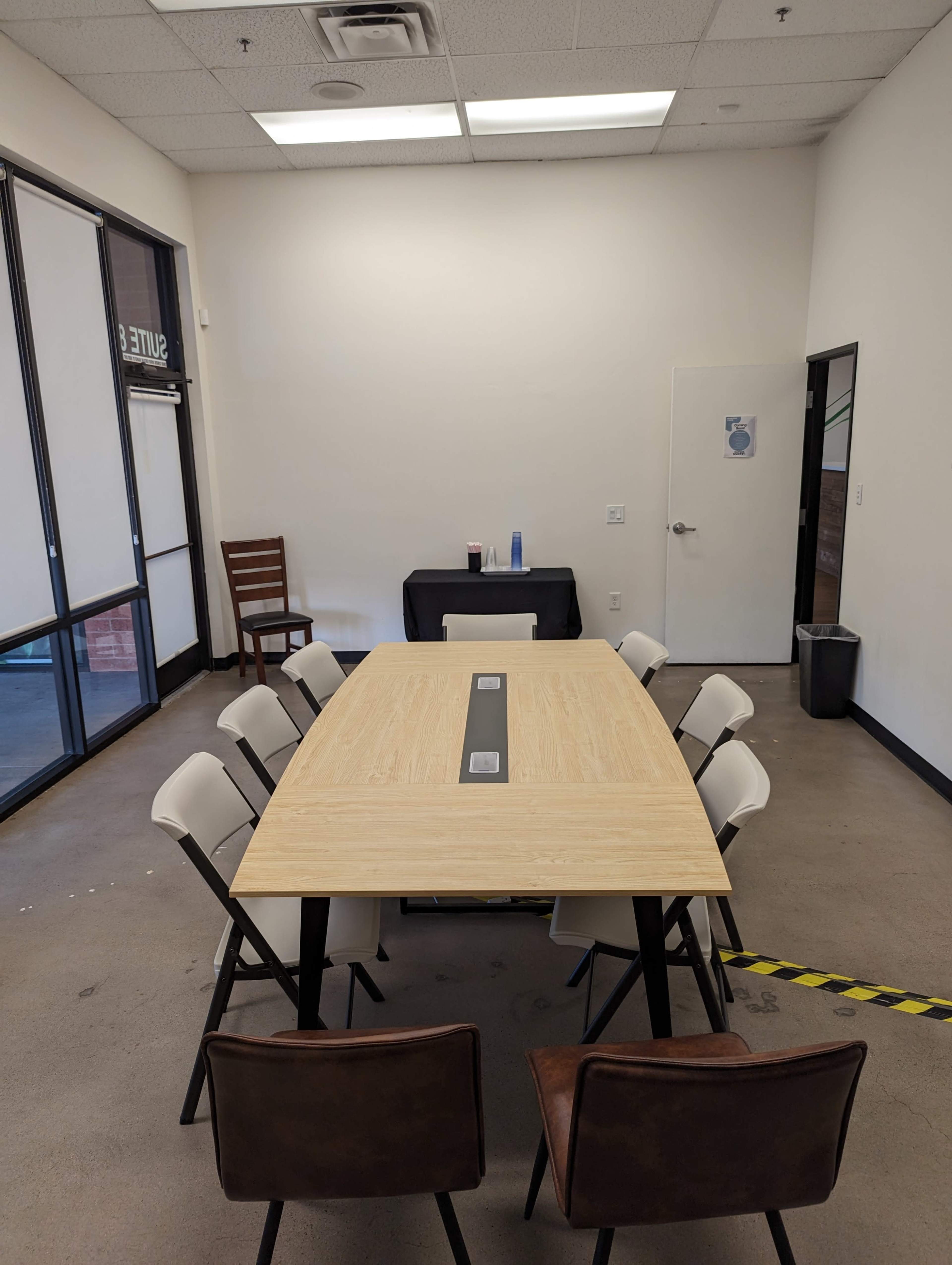 A rectangular conference table with eight chairs surrounds it in a plain meeting room, featuring a white wall and a door.