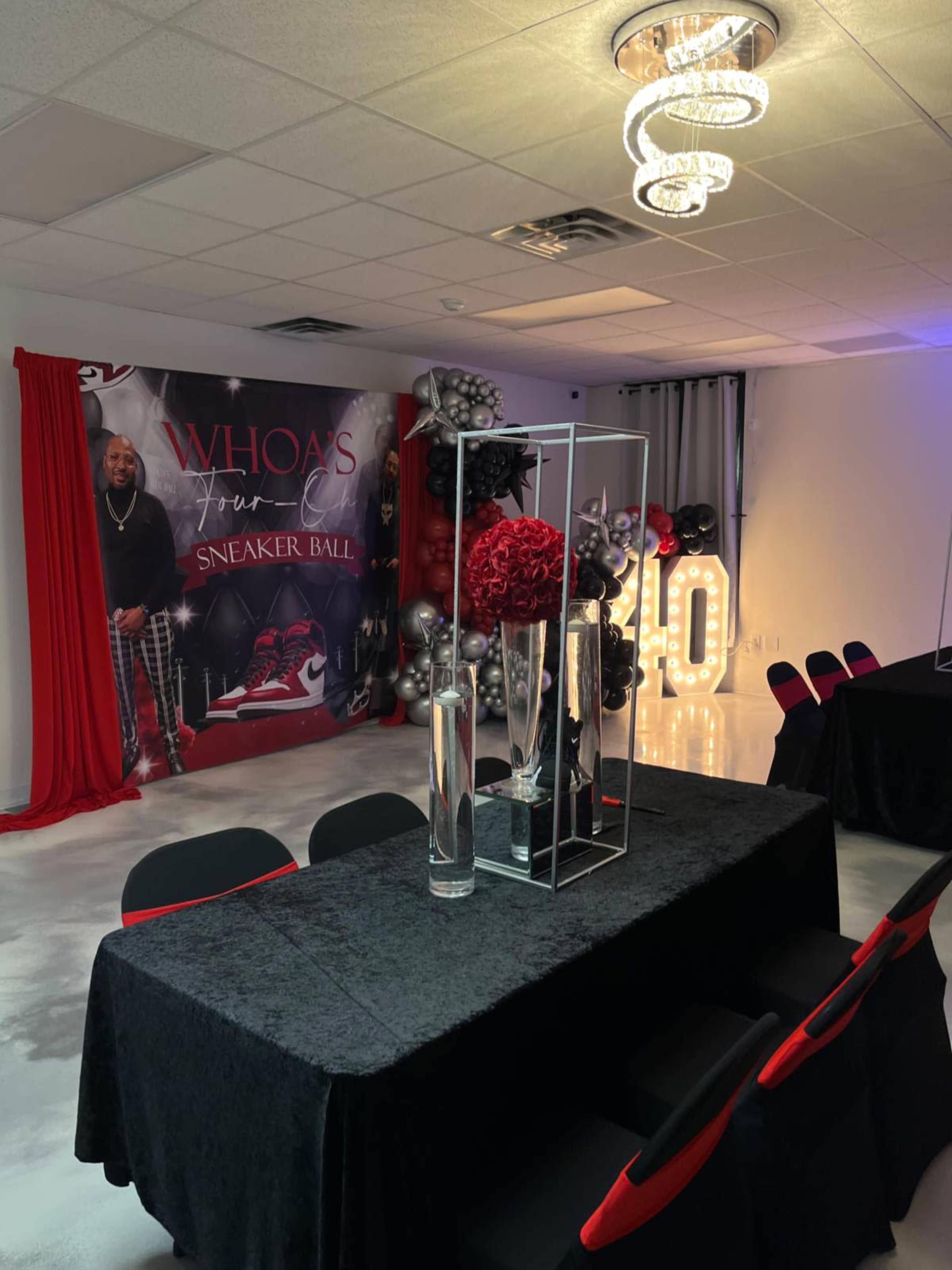 The image shows a decorated event space featuring a black table with red accents, a backdrop for a sneaker ball, and a display of floral arrangements within glass containers.
