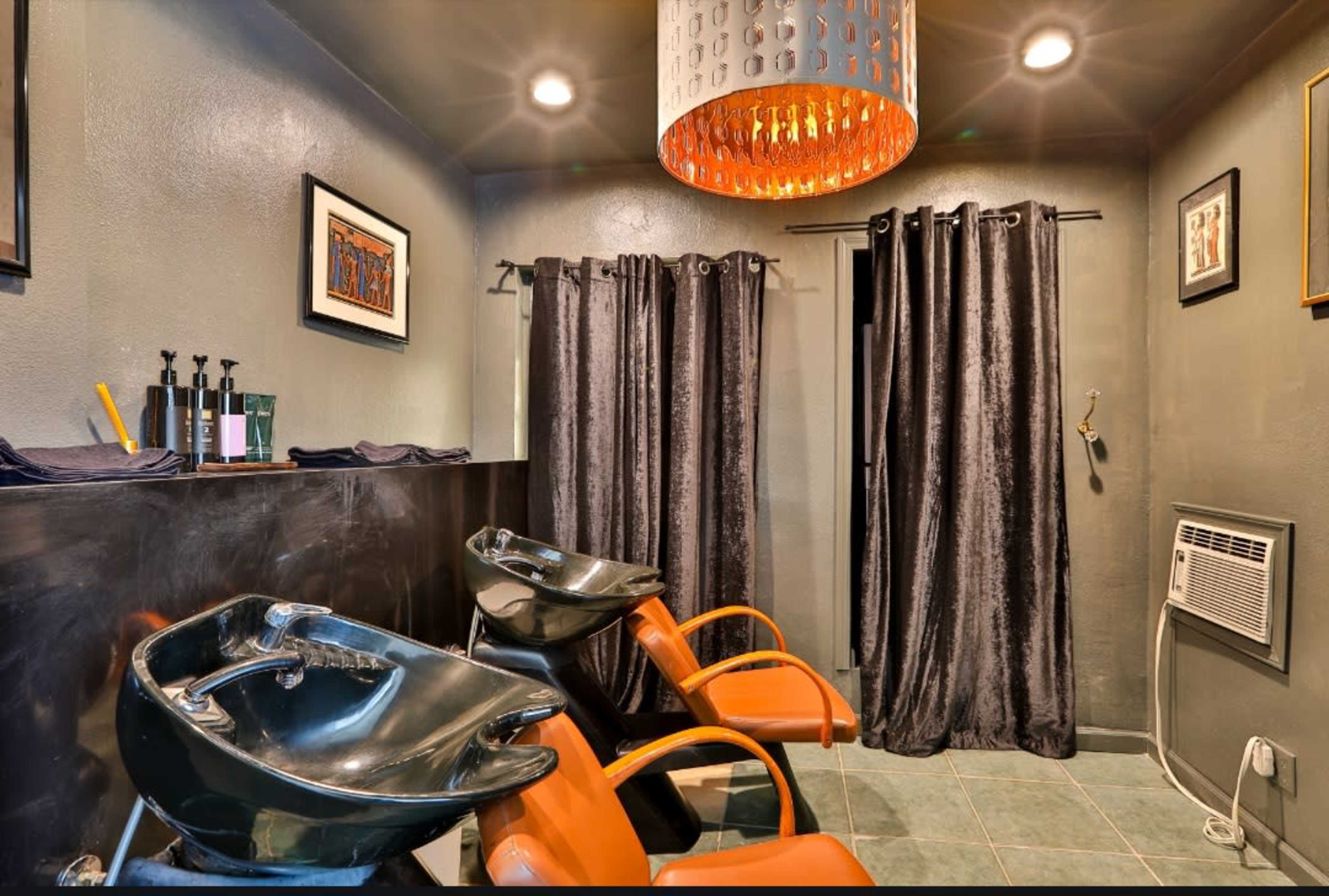 The image shows a hair salon interior with two black shampoo bowls, orange chairs, and gray curtains, illuminated by a decorative pendant light.