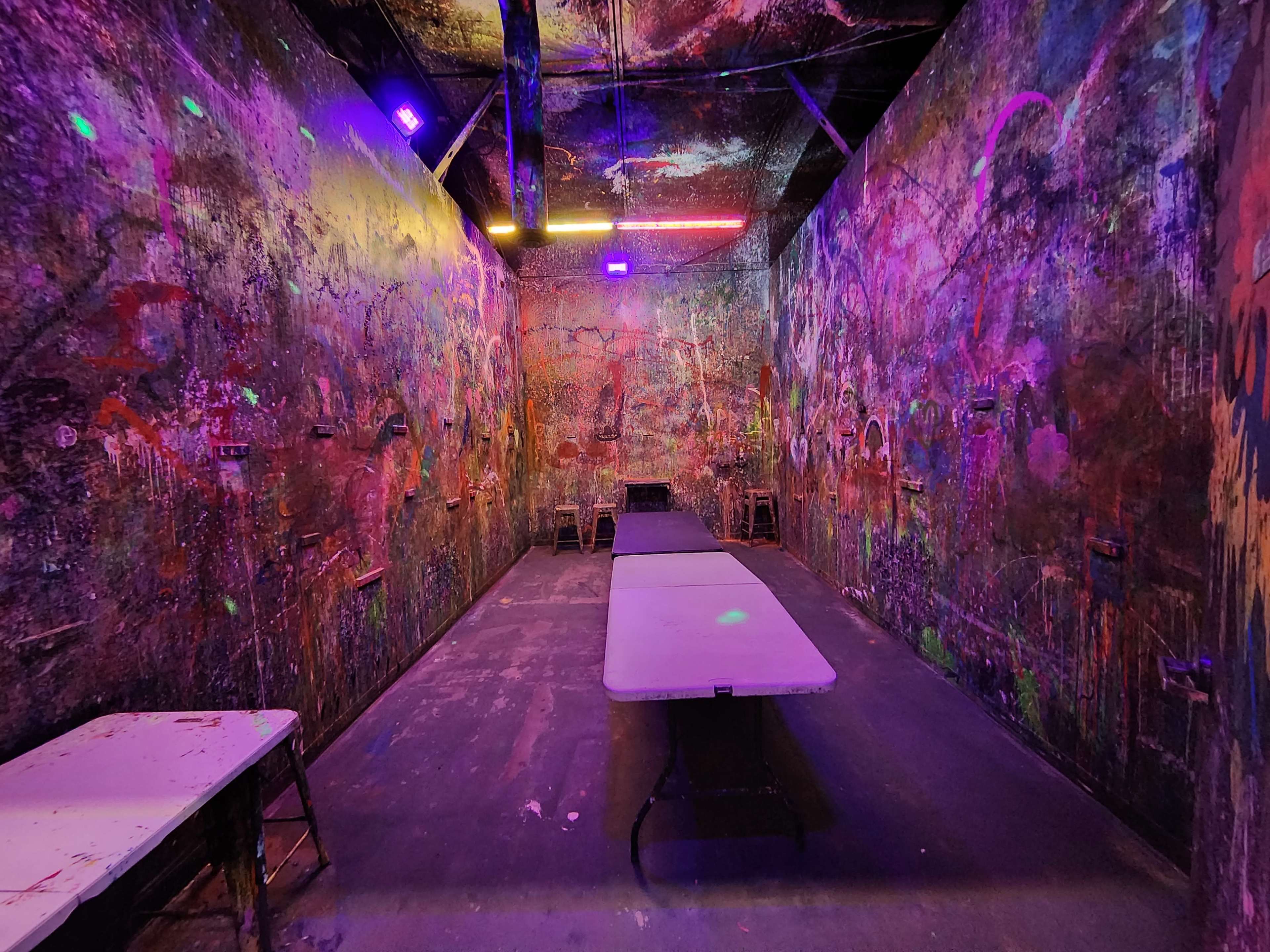The image shows a dimly lit room with colorful, artistically painted walls and a central folding table surrounded by several stacked chairs.