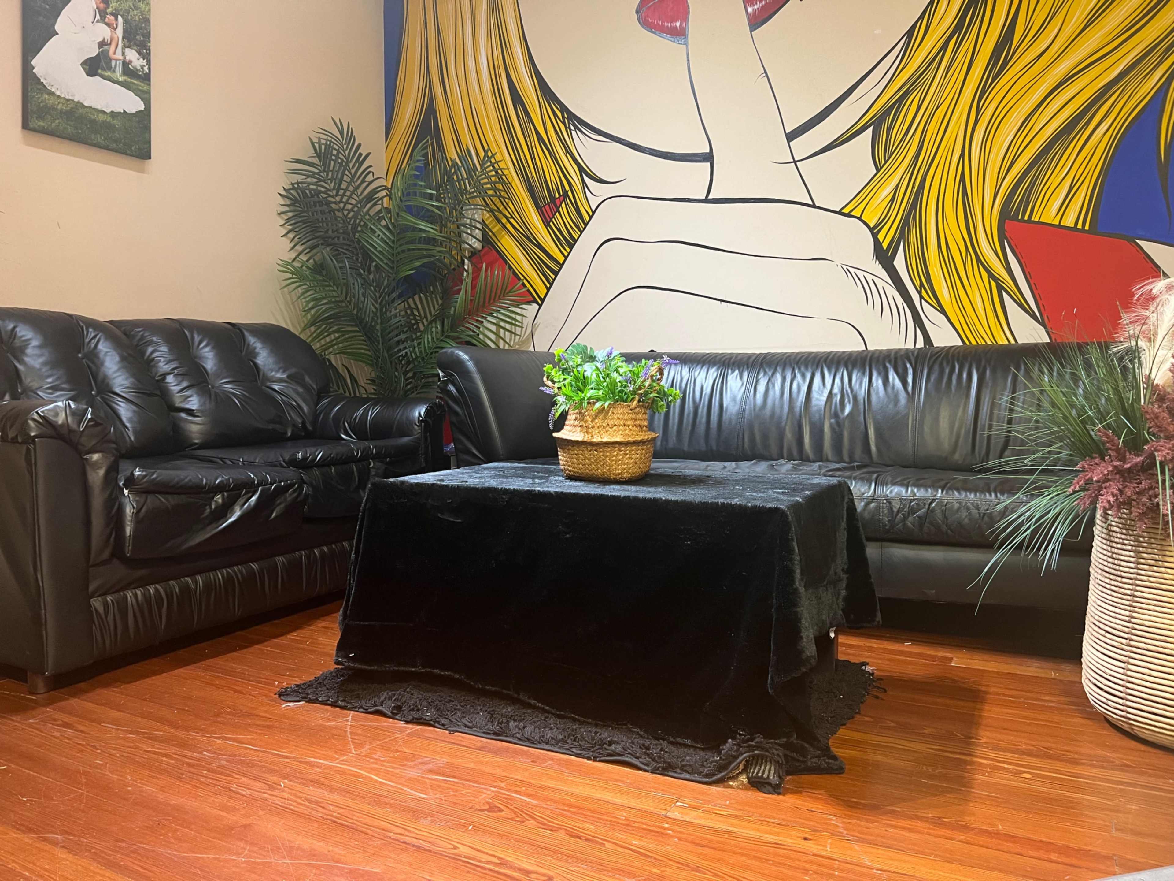The image shows a room with a black leather couch and a black table covered with a velvet cloth, surrounded by decorative plants and a vibrant mural on the wall.