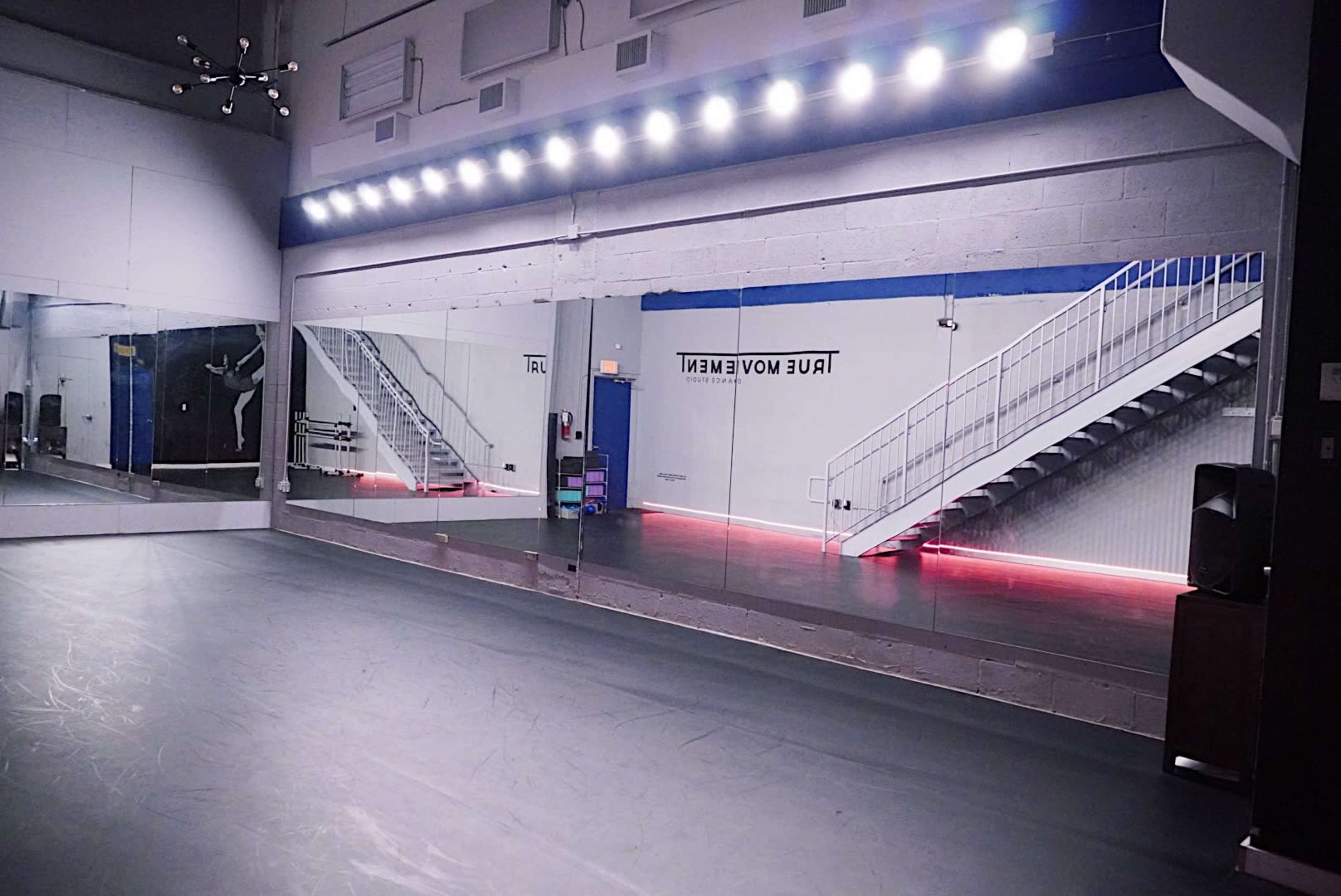 The image shows a dance studio with large mirrors, a staircase in the background, and LED lighting along the floor.