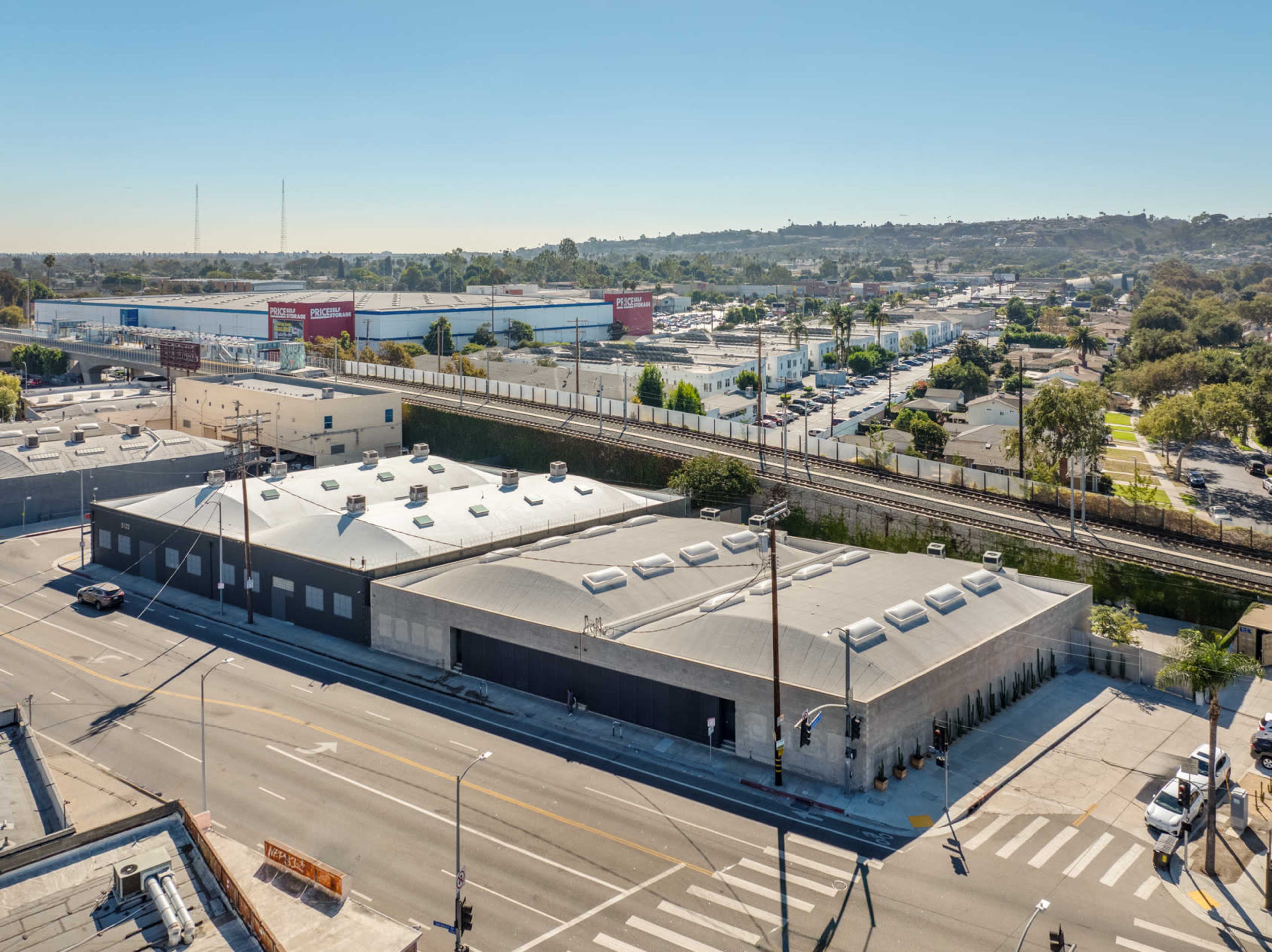 The image shows a large, modern building at the intersection of two streets, with a railway line and a series of warehouses in the background.