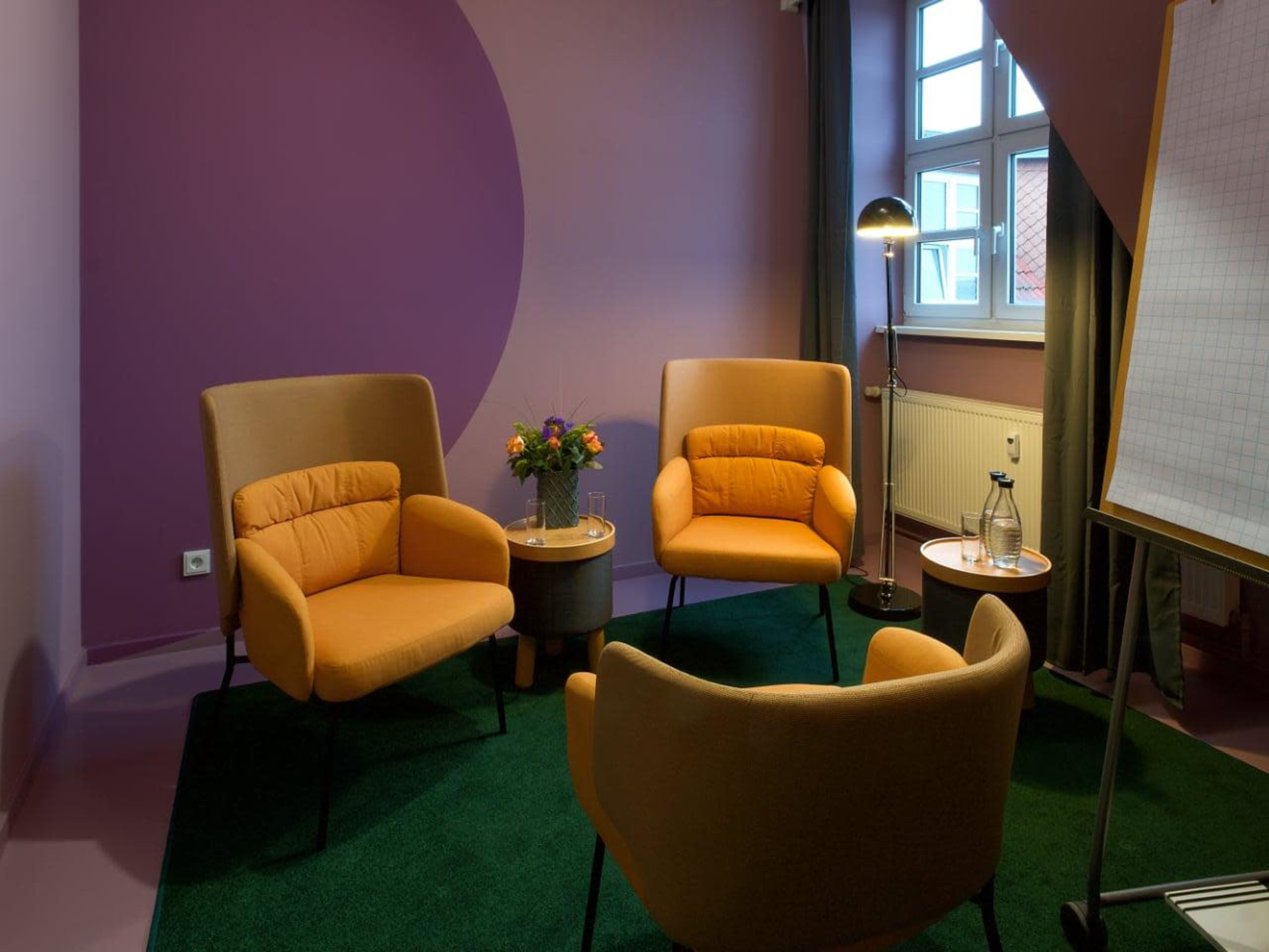 A modern meeting room with three orange chairs, a small round table, a lamp, and a whiteboard against a purple and pink wall.