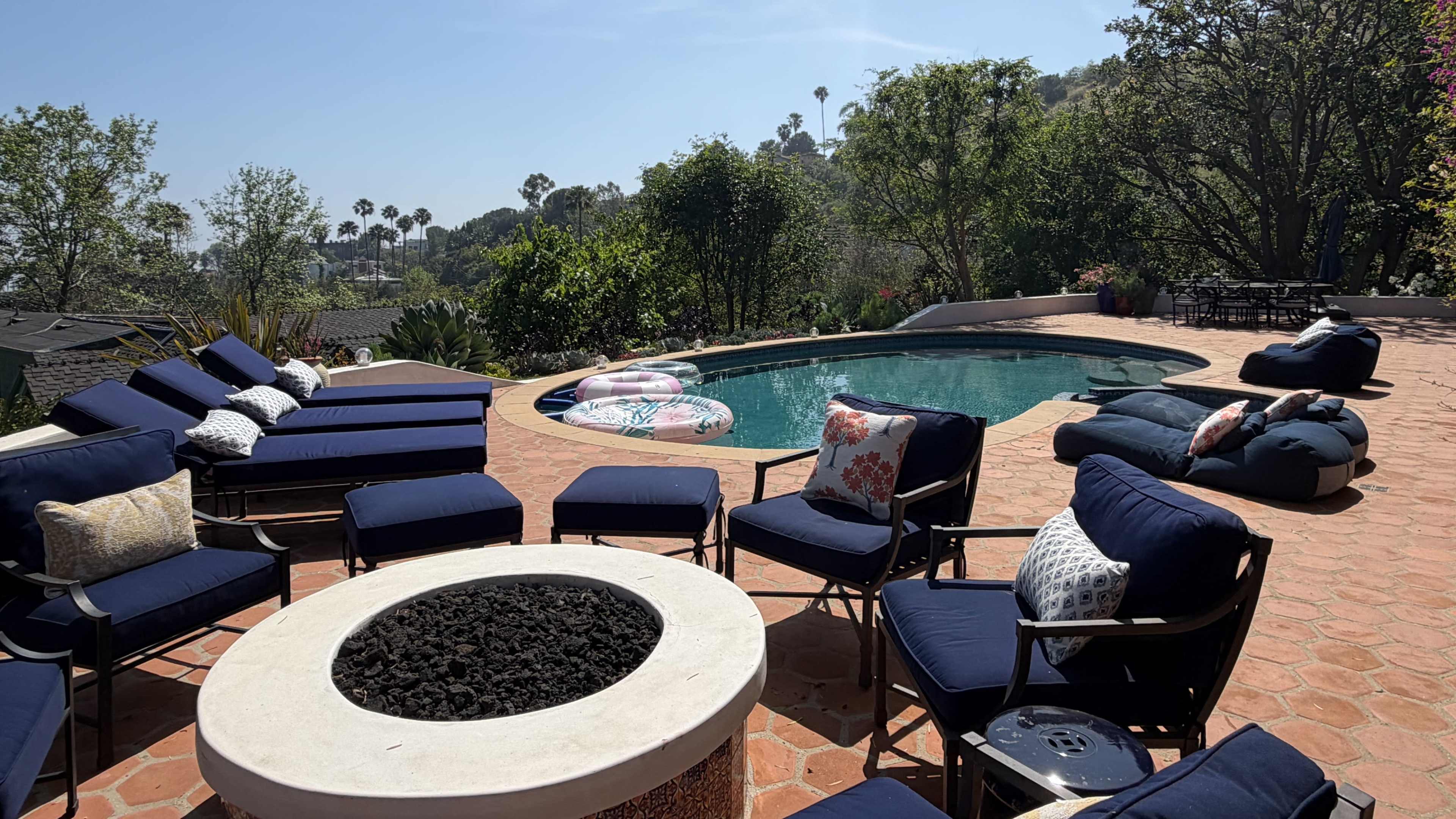 The image shows a backyard area with a pool, lounge chairs arranged around a fire pit, and inflatable pool floats nearby, surrounded by greenery and a clear blue sky.