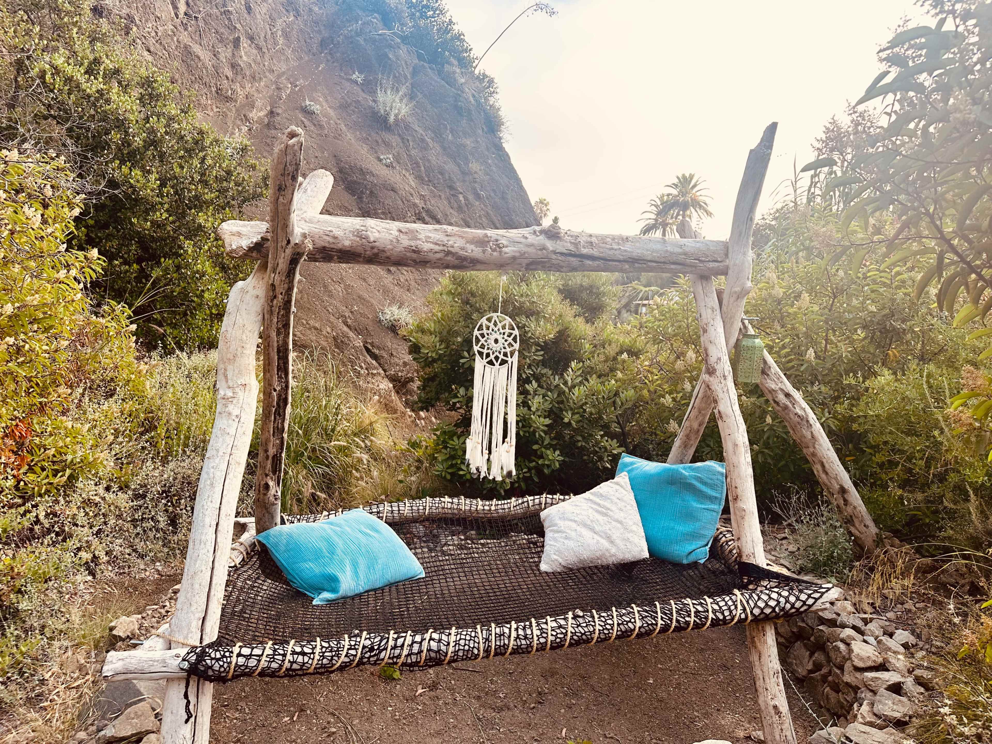 A rustic swing made of wooden logs hangs in a natural setting, adorned with a dreamcatcher and two blue pillows.