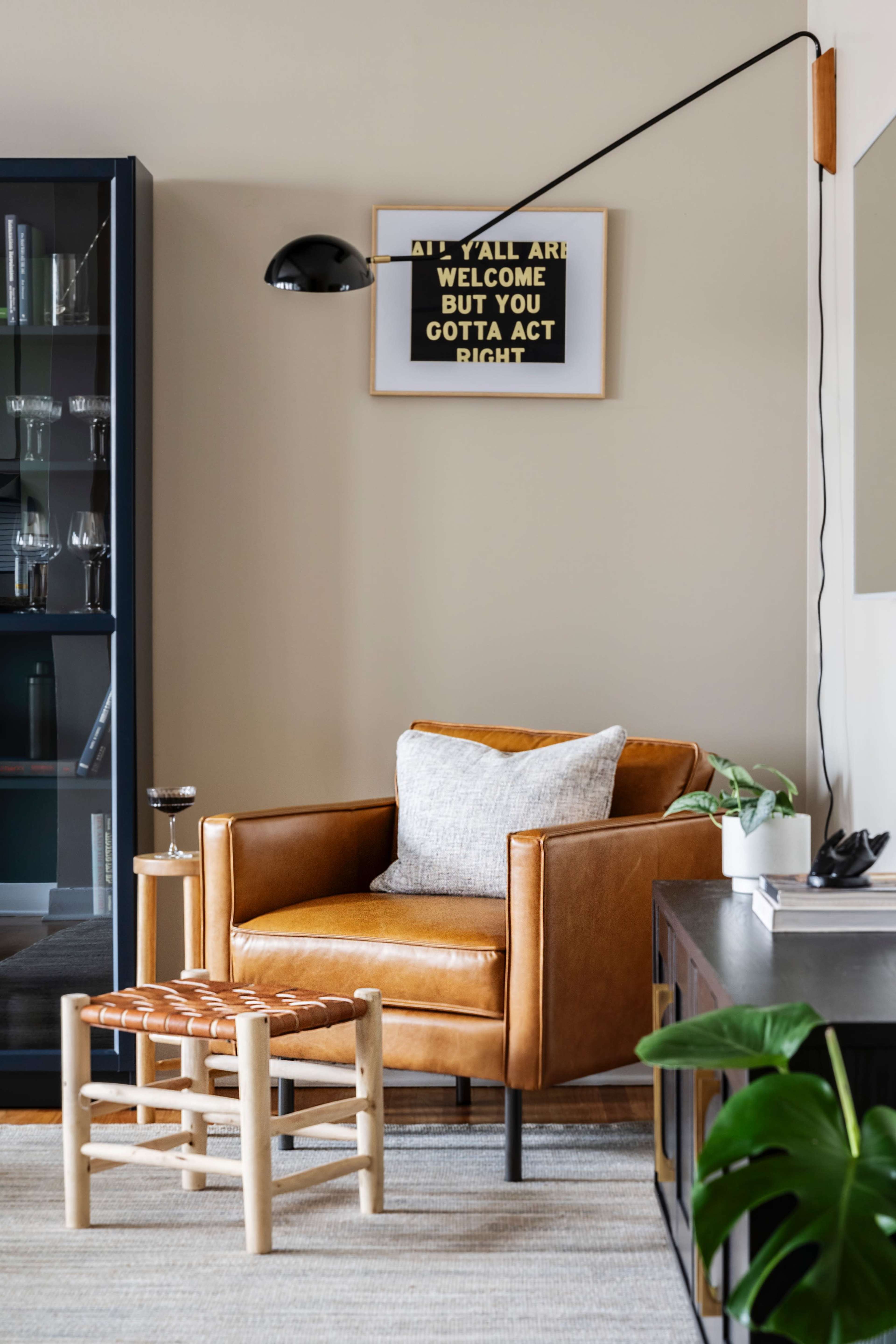 A brown leather armchair with a cushion sits next to a small wooden stool, while a wall lamp illuminates a framed sign that reads, "ALL Y'ALL ARE WELCOME BUT YOU GOTTA ACT RIGHT."