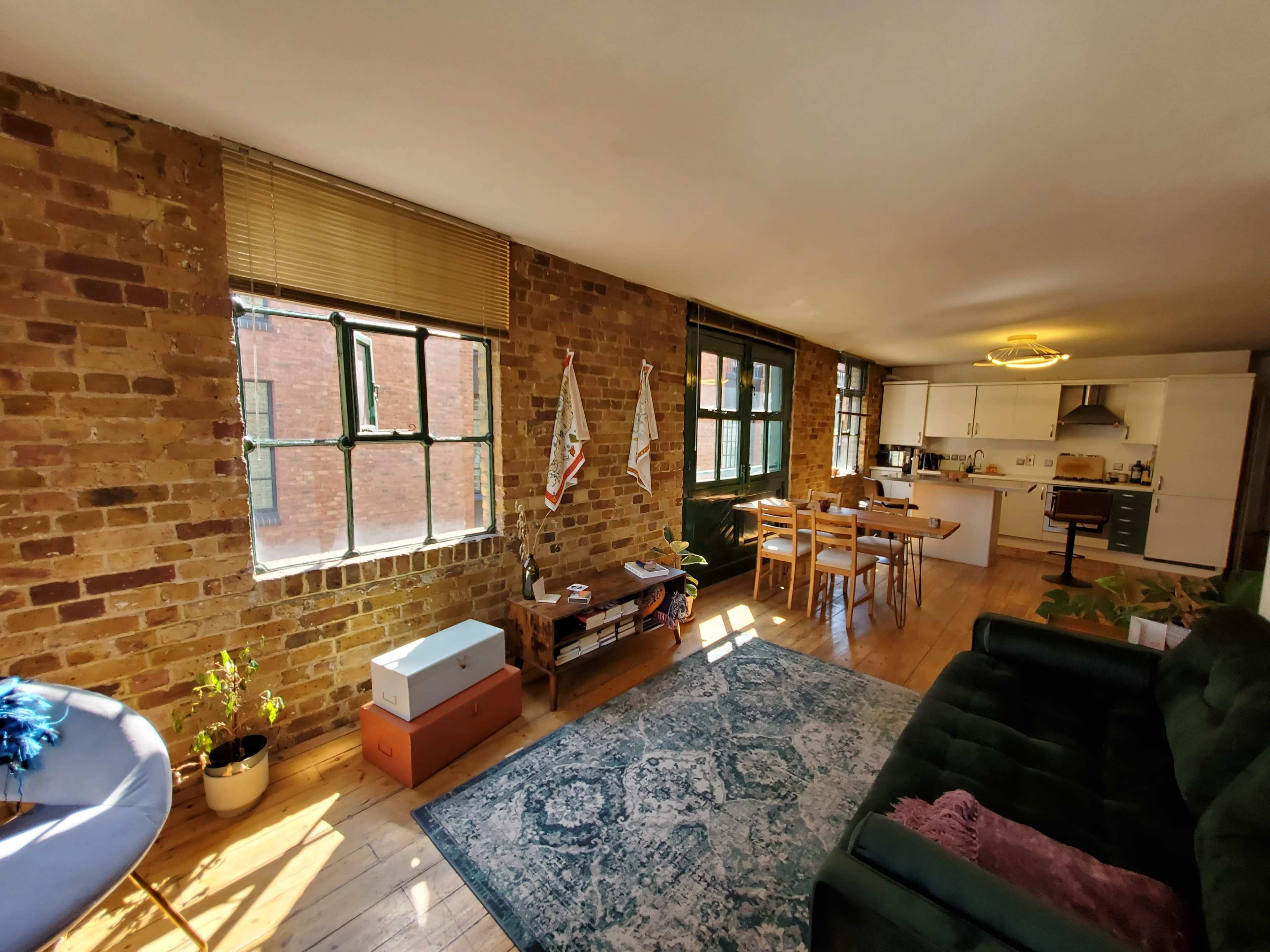 A spacious living area with exposed brick walls, large windows, a dark sofa, a dining table, and a kitchen in the background.
