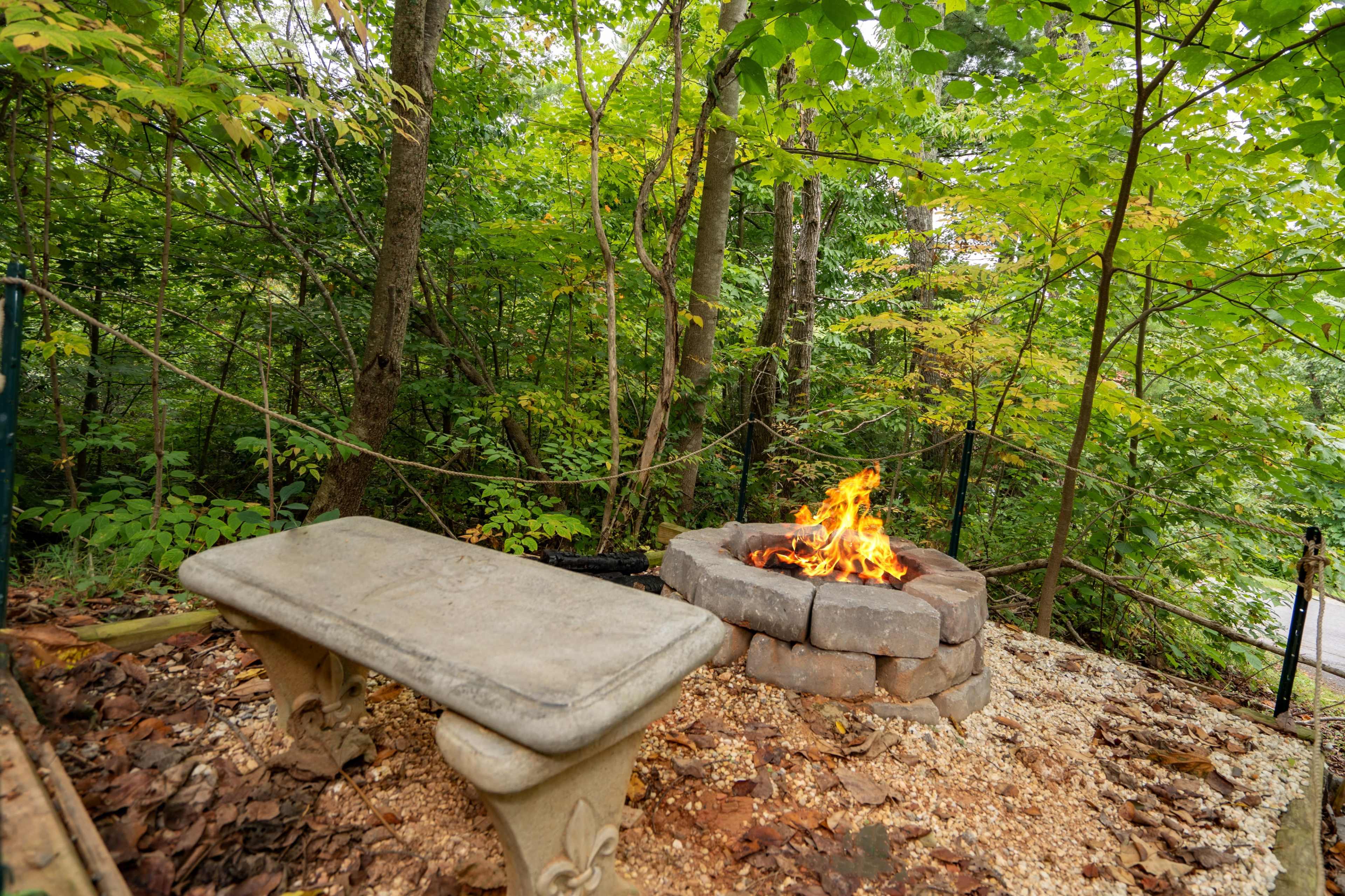 A stone fire pit surrounded by a concrete bench is nestled in a wooded area with green trees and fallen leaves.