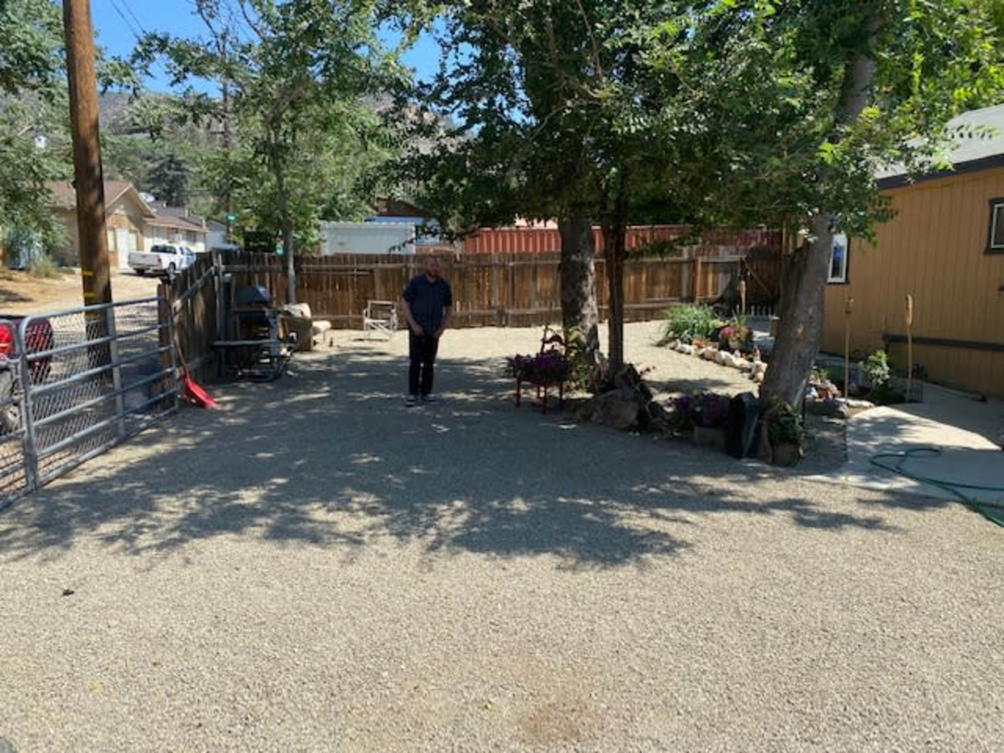 A person stands in a gravel backyard surrounded by trees and a wooden fence.