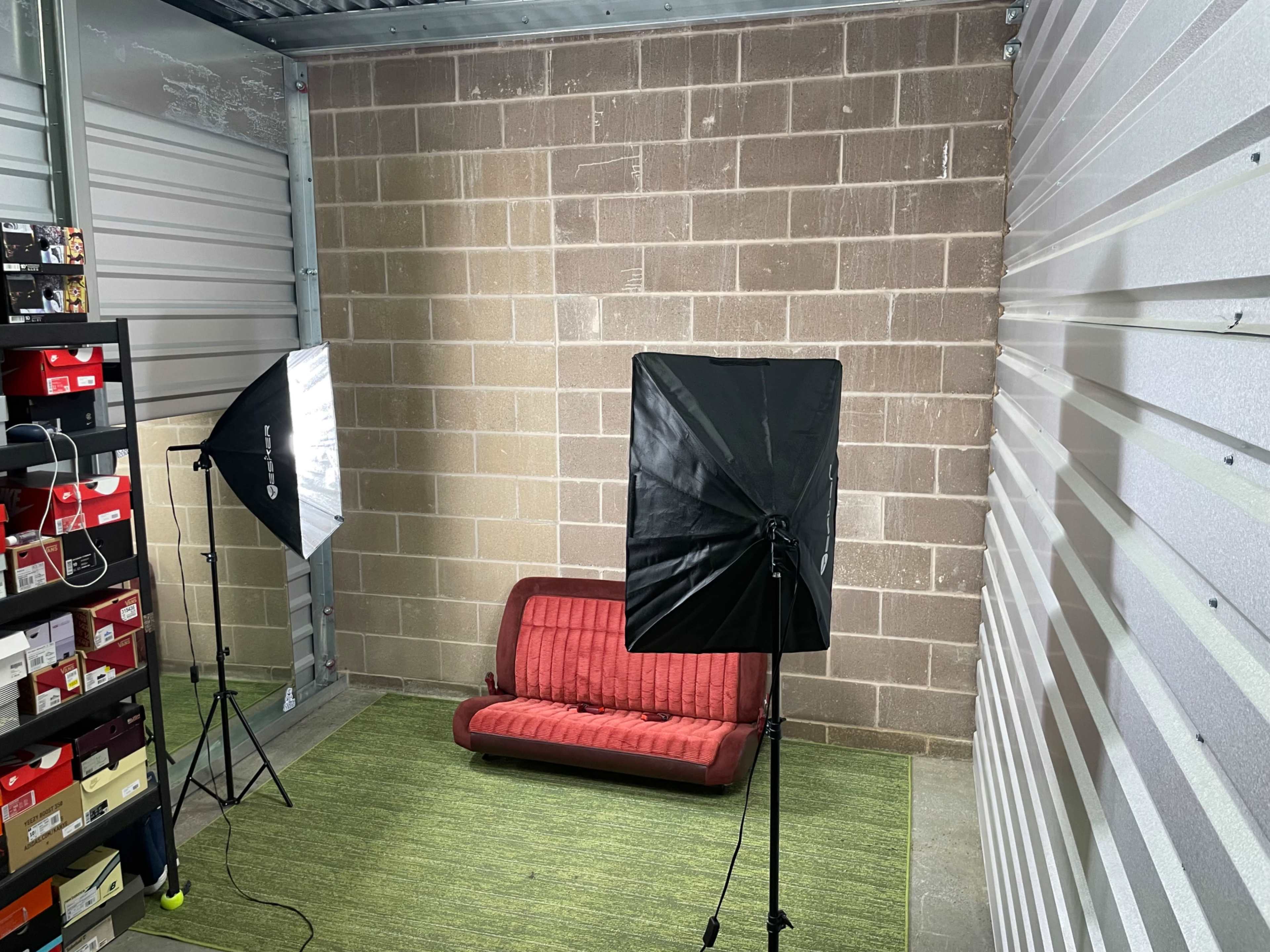 The image shows a small storage unit featuring a red couch, two softbox lights, and shelves filled with boxes along one wall.