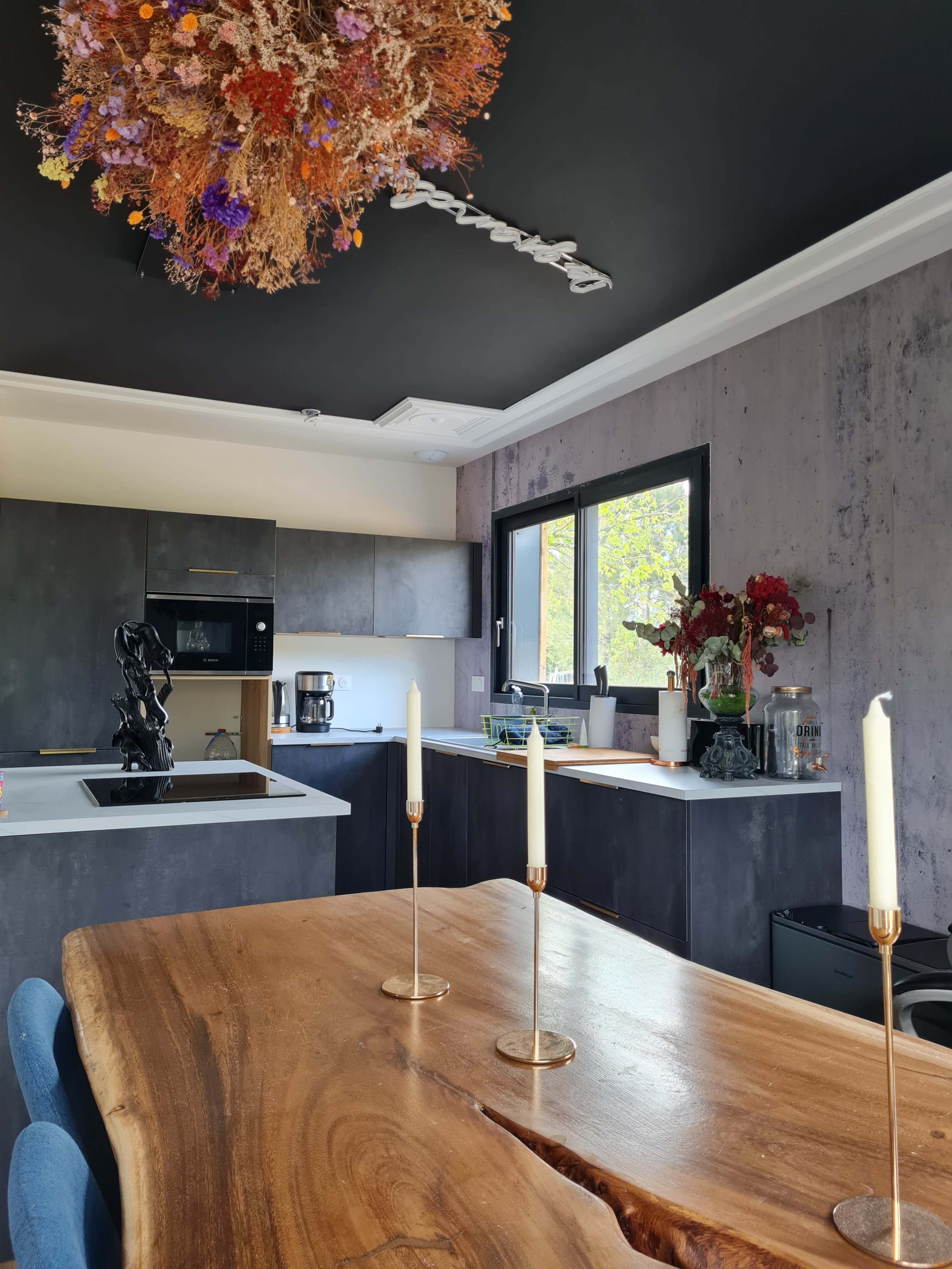A modern kitchen with dark cabinetry, a central white island, and a large wooden dining table featuring three candle holders, illuminated by a colorful hanging light fixture.