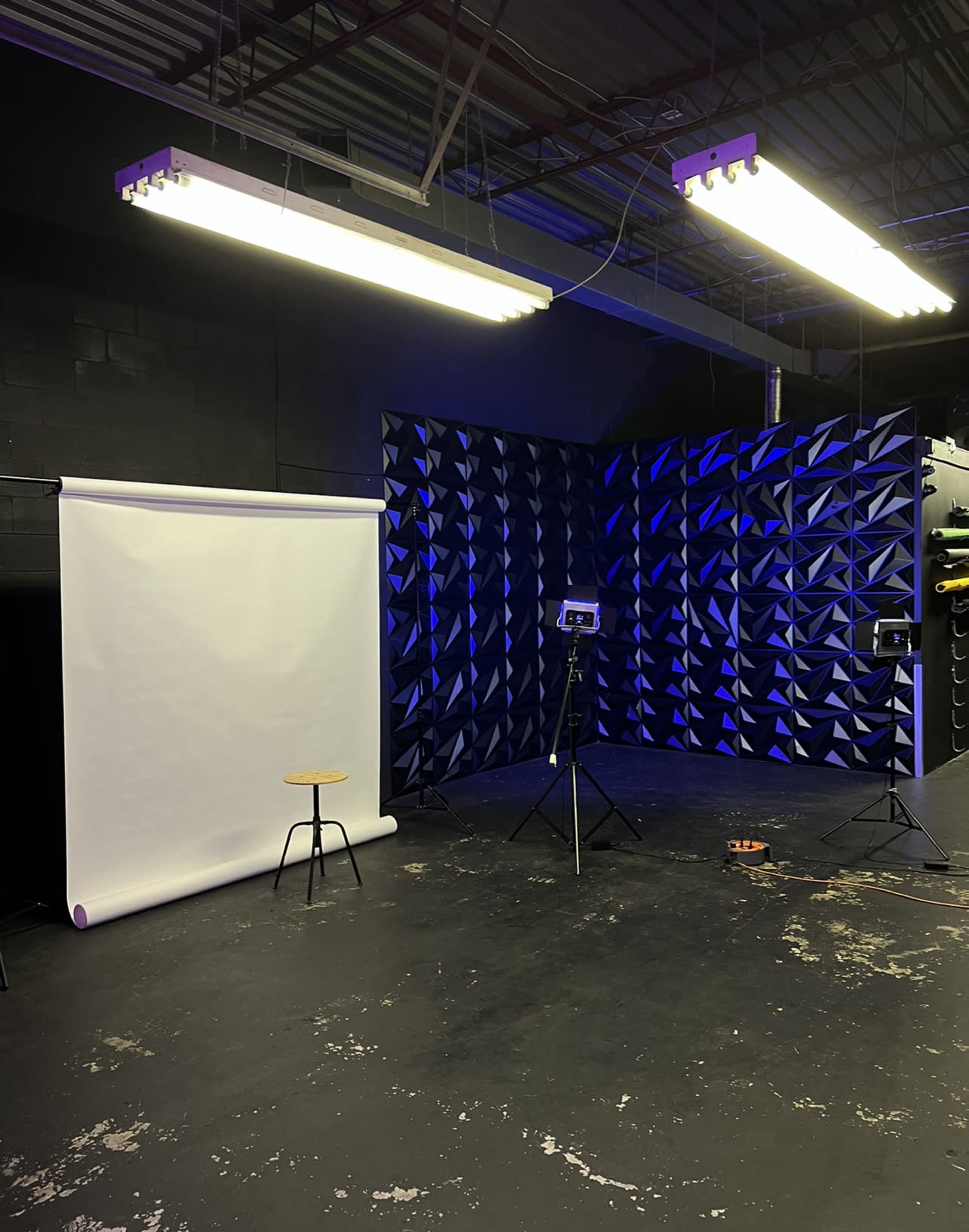 The image shows a photography studio with a white backdrop, a stool, and two lighting setups against a textured blue wall.