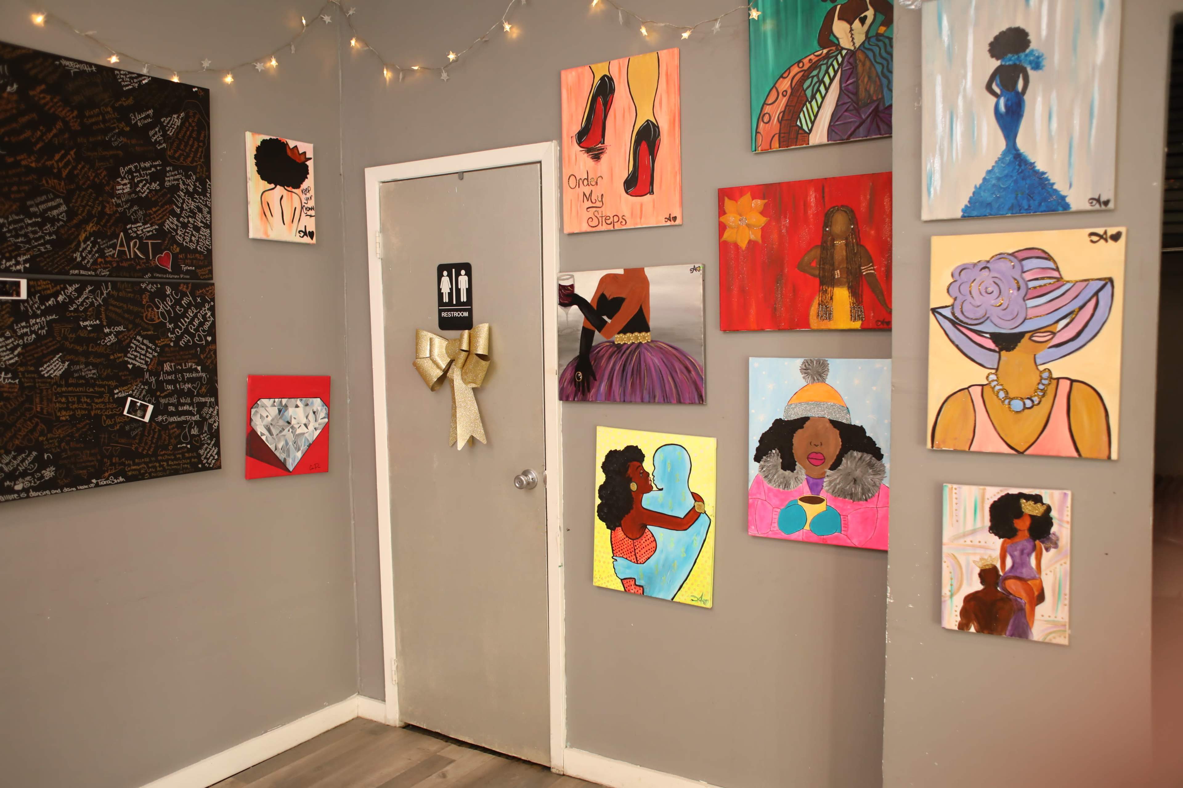 A gallery wall decorated with various colorful paintings featuring women and fashion, alongside a door marked with restroom symbols.