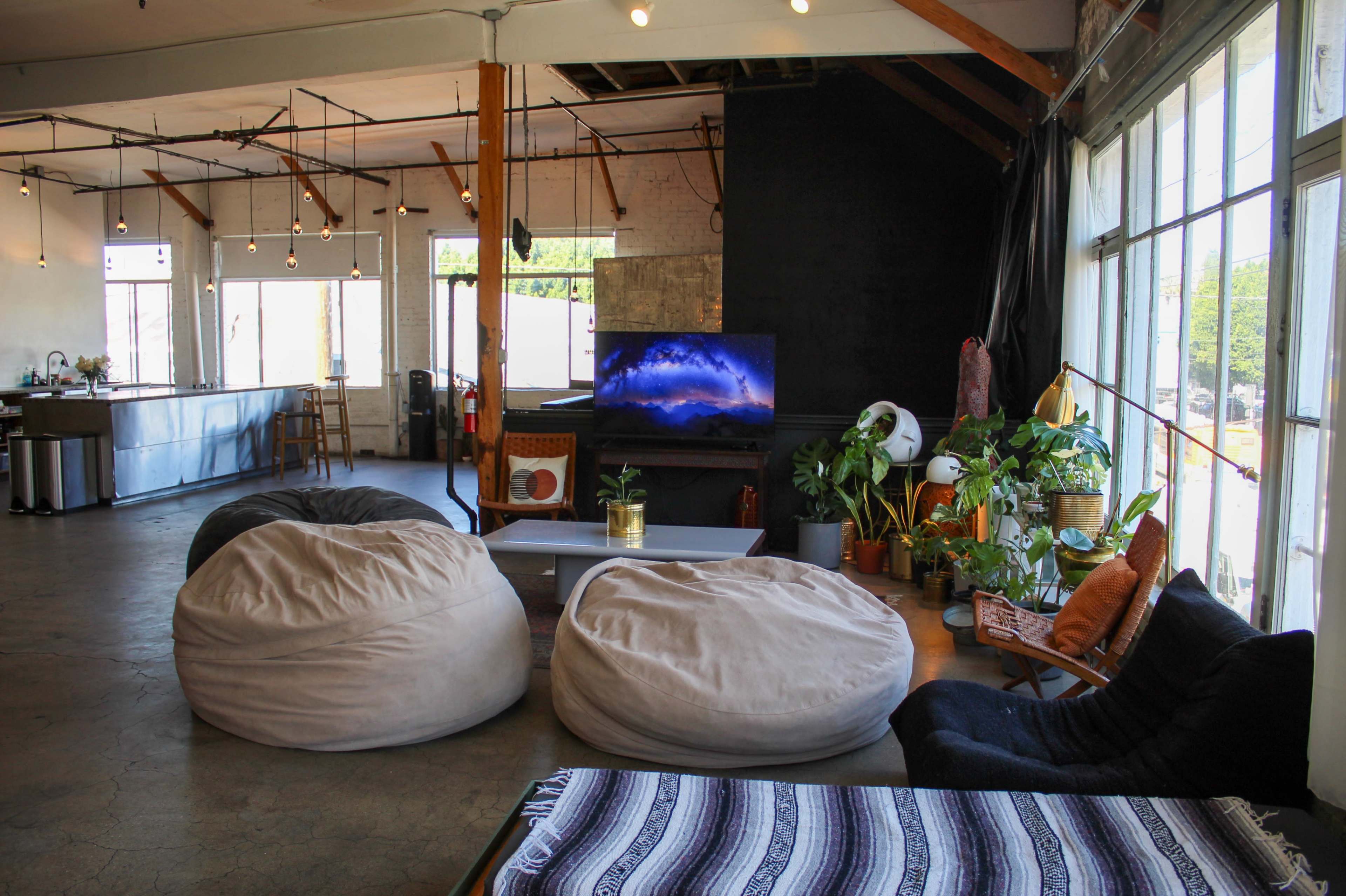 The image shows a spacious, modern loft with large windows, comfortable bean bag chairs, a television on display, and various indoor plants arranged around a low coffee table.