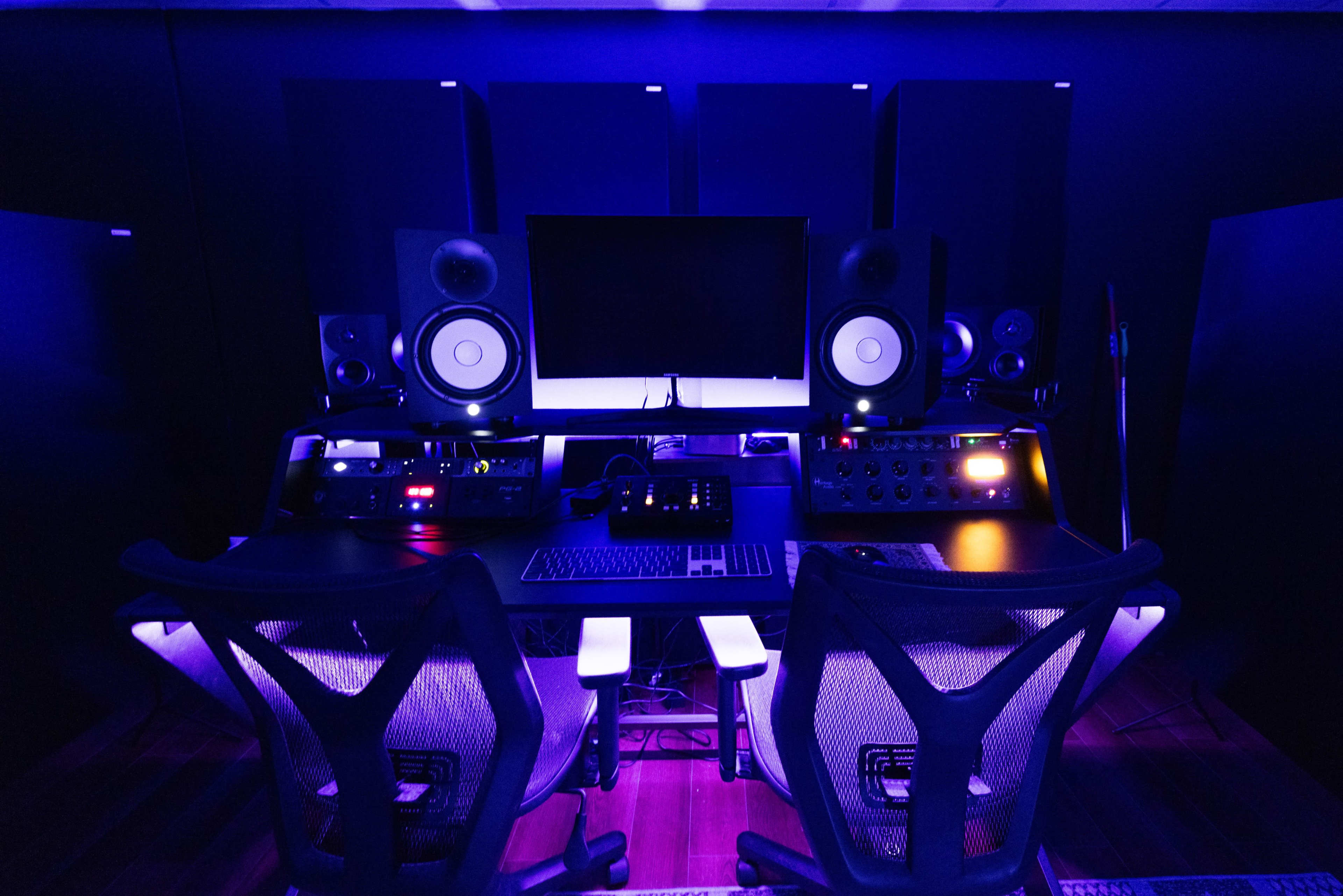 A music production studio featuring a digital audio workstation setup with two ergonomic chairs and multiple speakers arranged around a central desk.