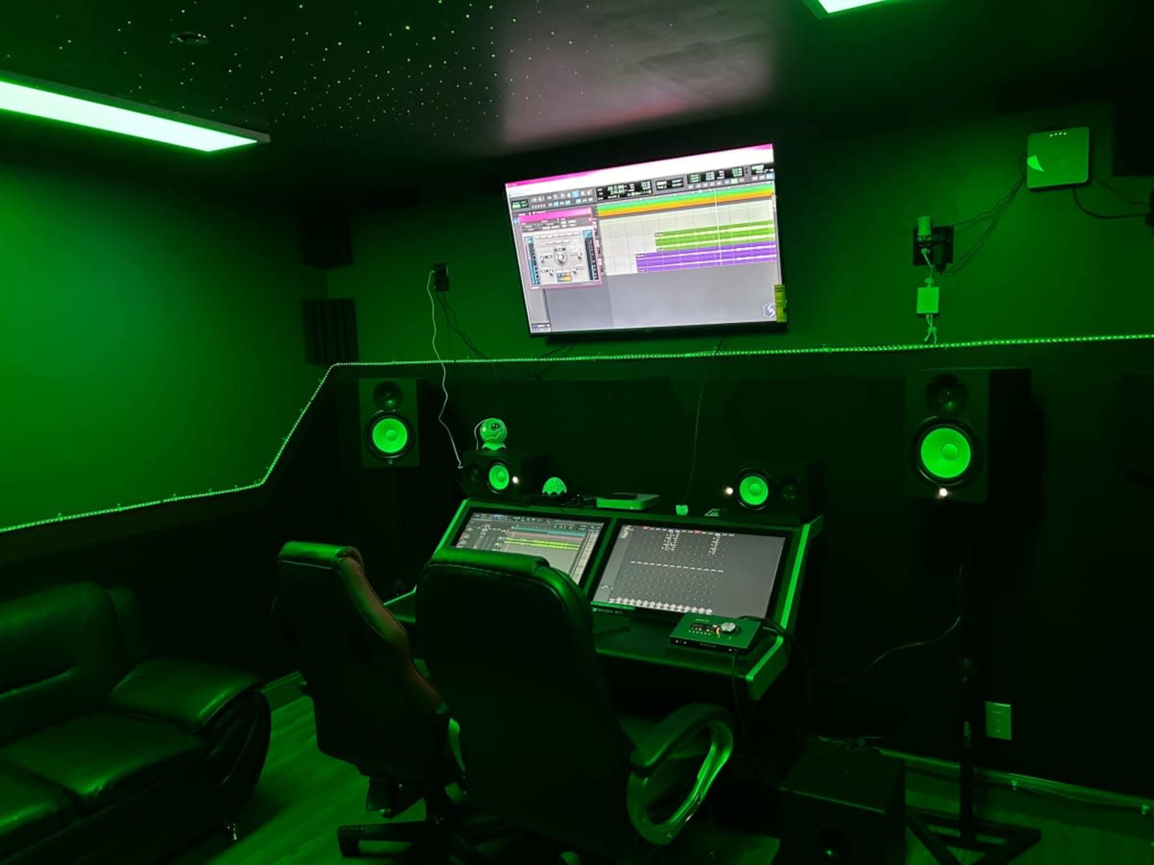 The image shows a dimly lit music studio featuring a desk with audio equipment, large speakers, and a wall-mounted monitor displaying software tracks.