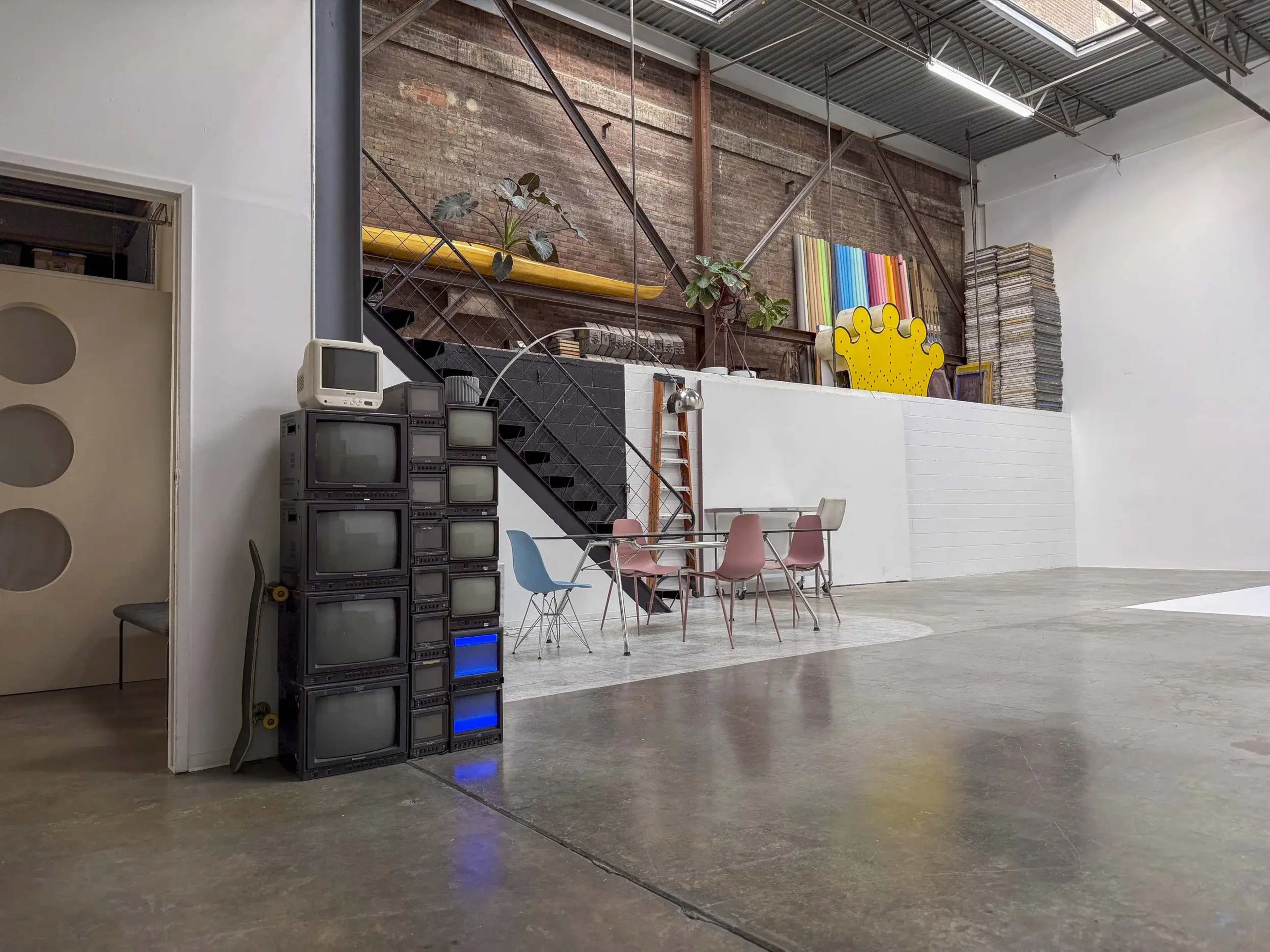 The image shows a spacious industrial-style interior with a staircase, a ladder, a colorful wall hanging, a set of pastel chairs around a table, and a stack of vintage televisions.