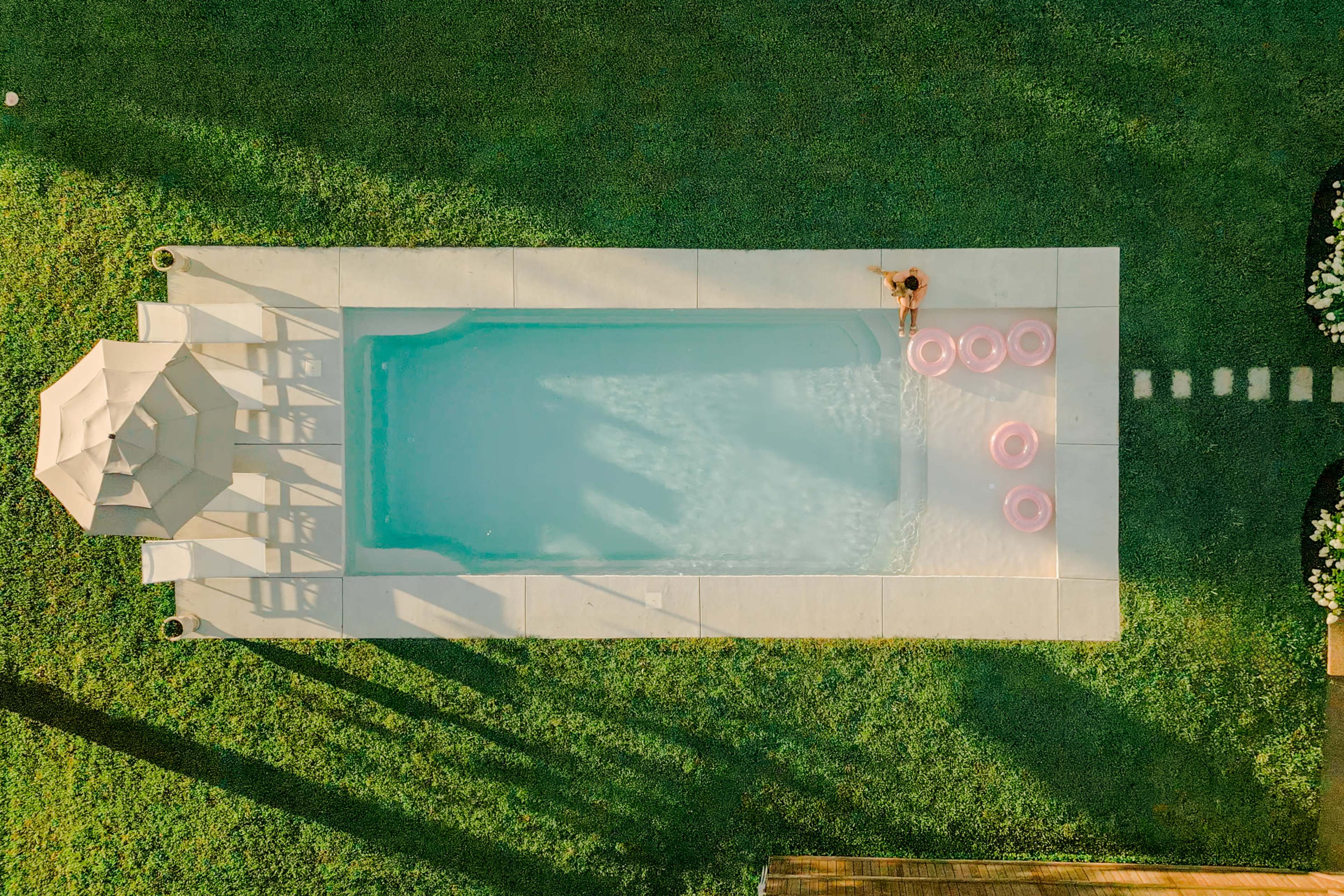 An aerial view shows a rectangular swimming pool surrounded by a neatly manicured lawn, with a gazebo nearby and floating pink pool floats in the water.