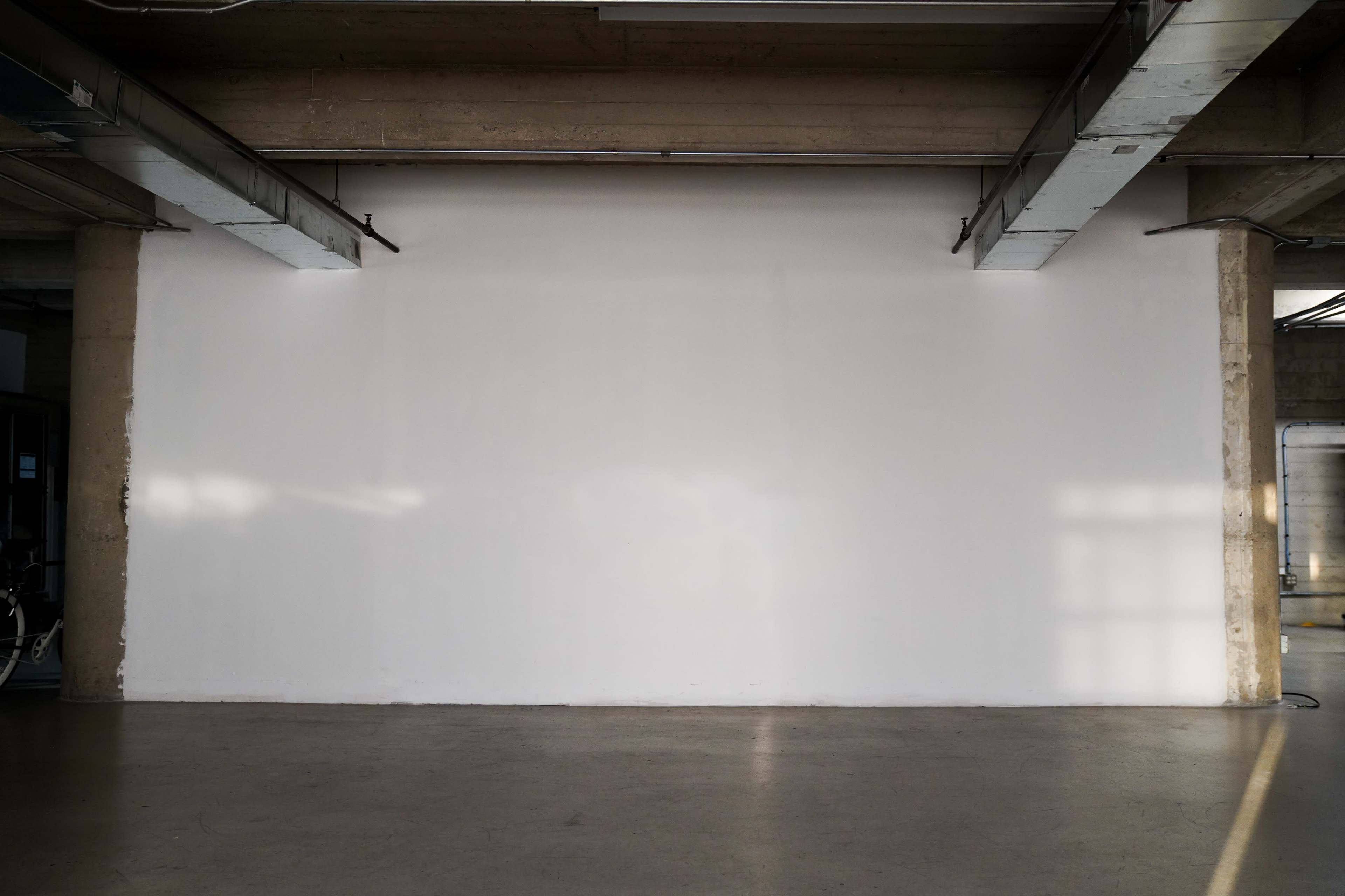 The image shows a large, empty white wall in an industrial-style room with concrete ceilings and floors.