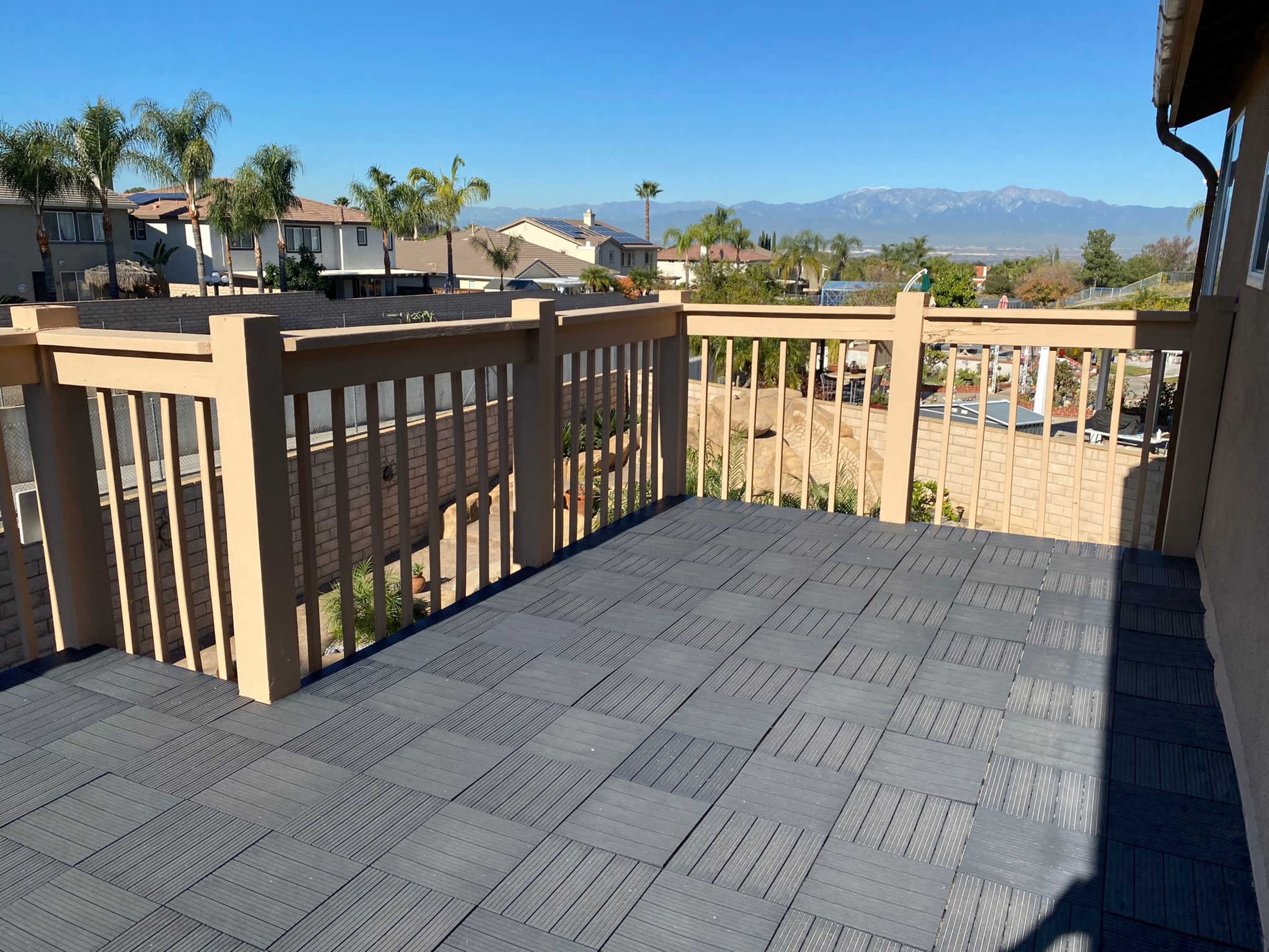 The image shows a wooden deck with interlocking tiles, enclosed by a beige railing, overlooking residential buildings and mountains in the distance under a clear sky.