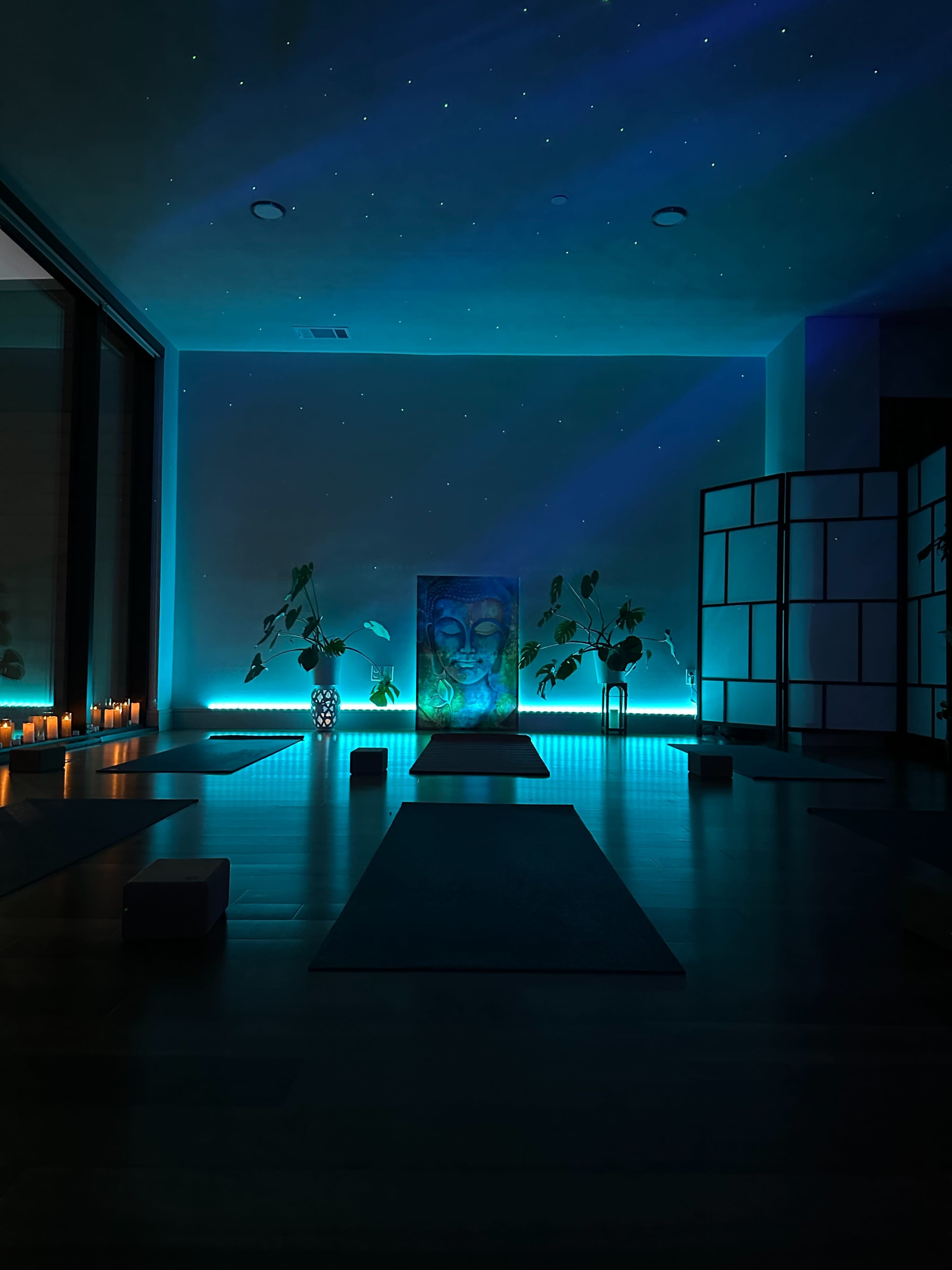 The image shows a serene yoga studio featuring a central Buddha painting, illuminated by soft blue lighting, surrounded by plants and candles.