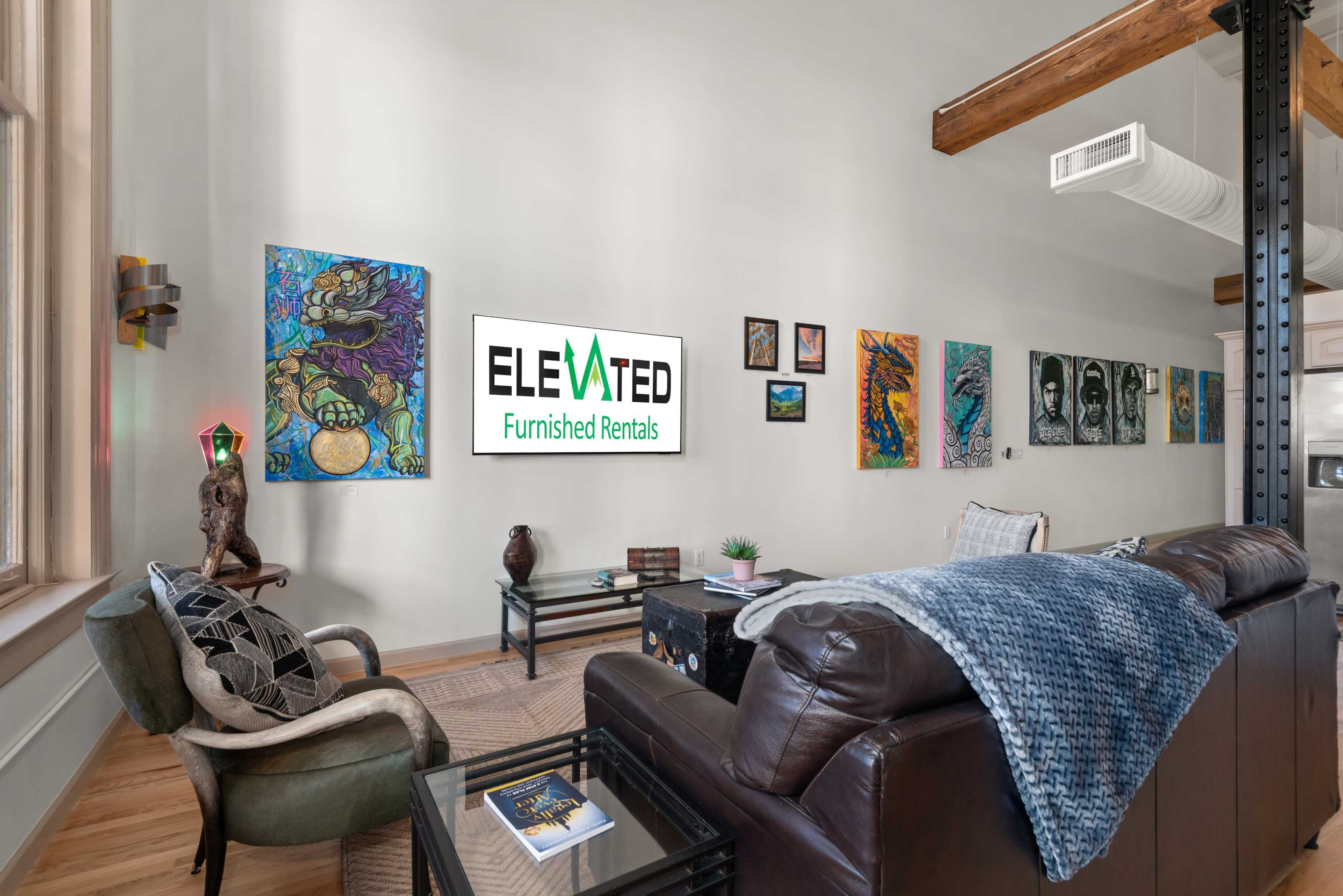 A modern living room features a leather sofa, a cozy chair, and diverse artwork on the walls, with a television displaying "ELEVATED Furnished Rentals."