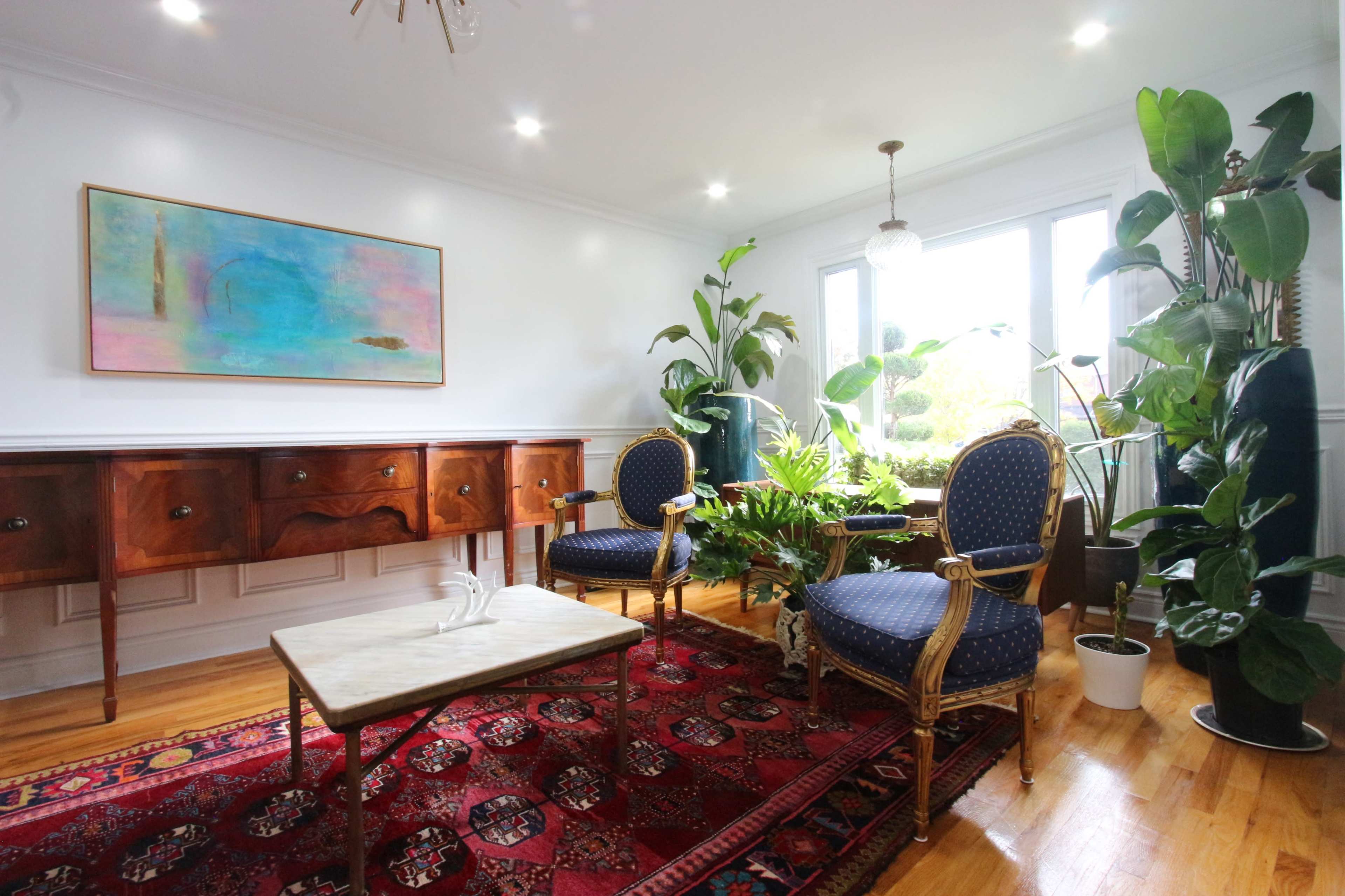 The image shows a well-lit living room with two blue upholstered chairs, a marble-top coffee table, a wooden sideboard, and several potted plants, all set against a colorful wall art piece.