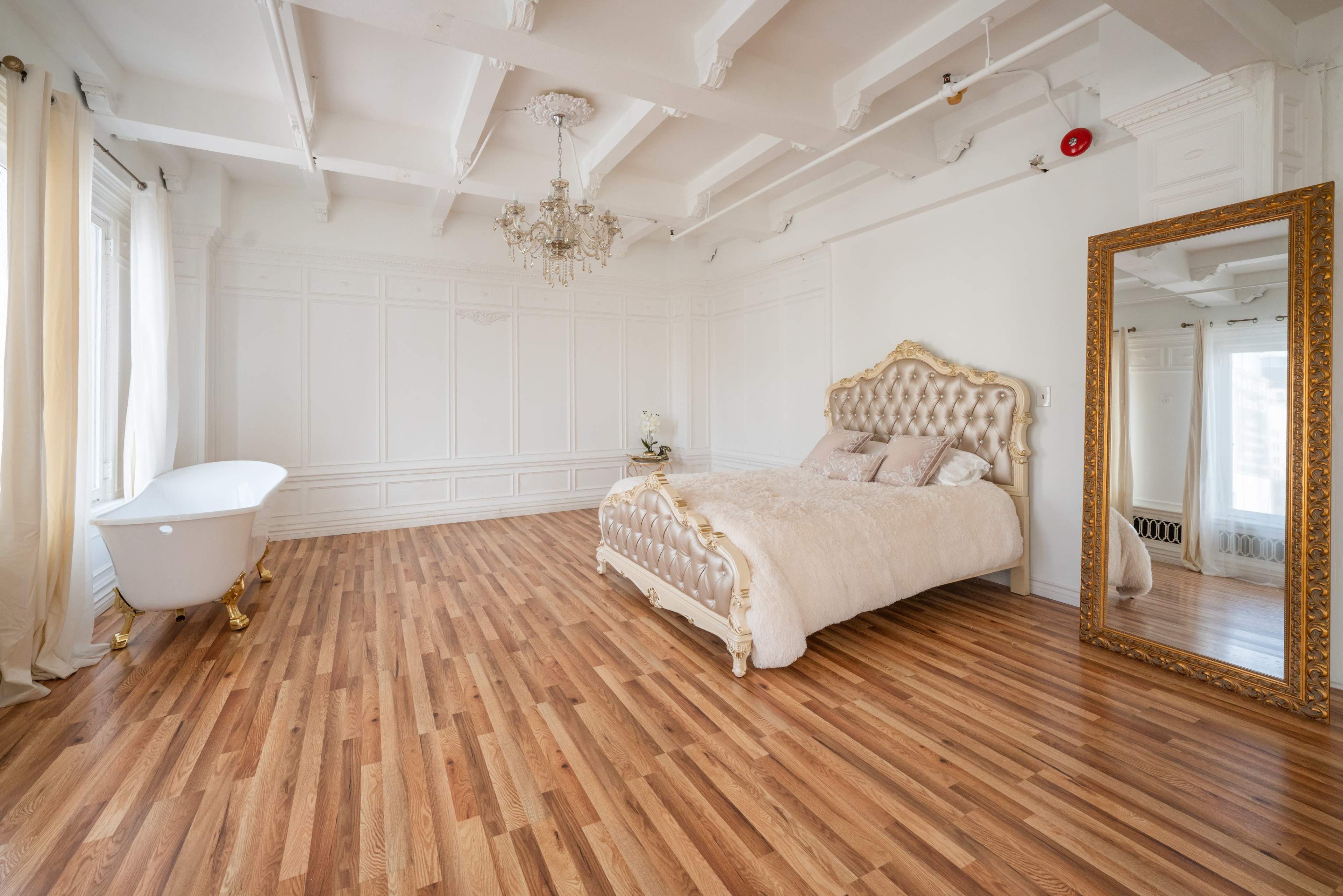 A spacious bedroom features a tufted gold bed, a freestanding bathtub, and a large ornate mirror, all set against white walls and wooden flooring.