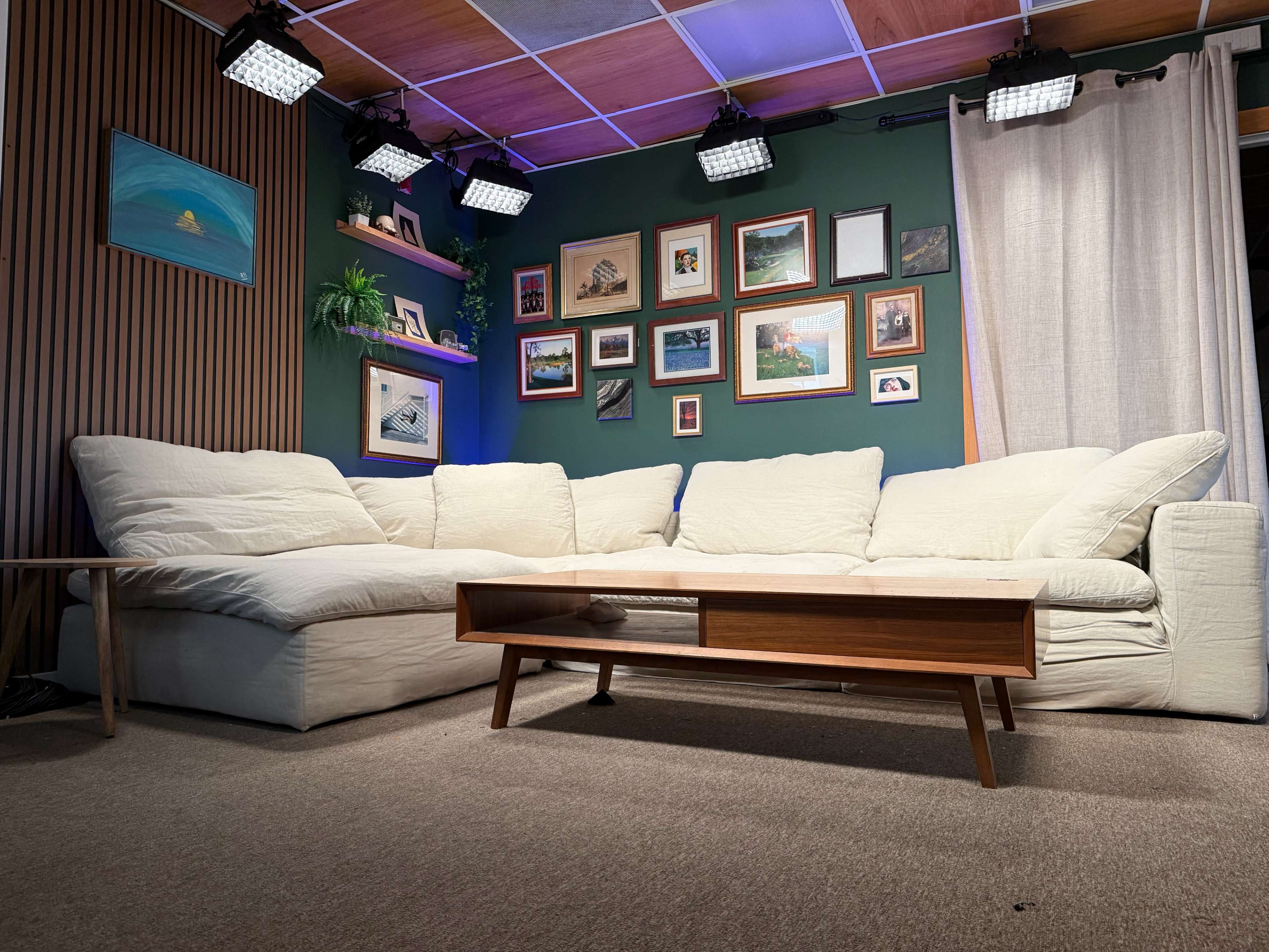 The image shows a cozy living room featuring a large white sectional sofa, a wooden coffee table, and several framed pictures on the walls, with bright lights above.