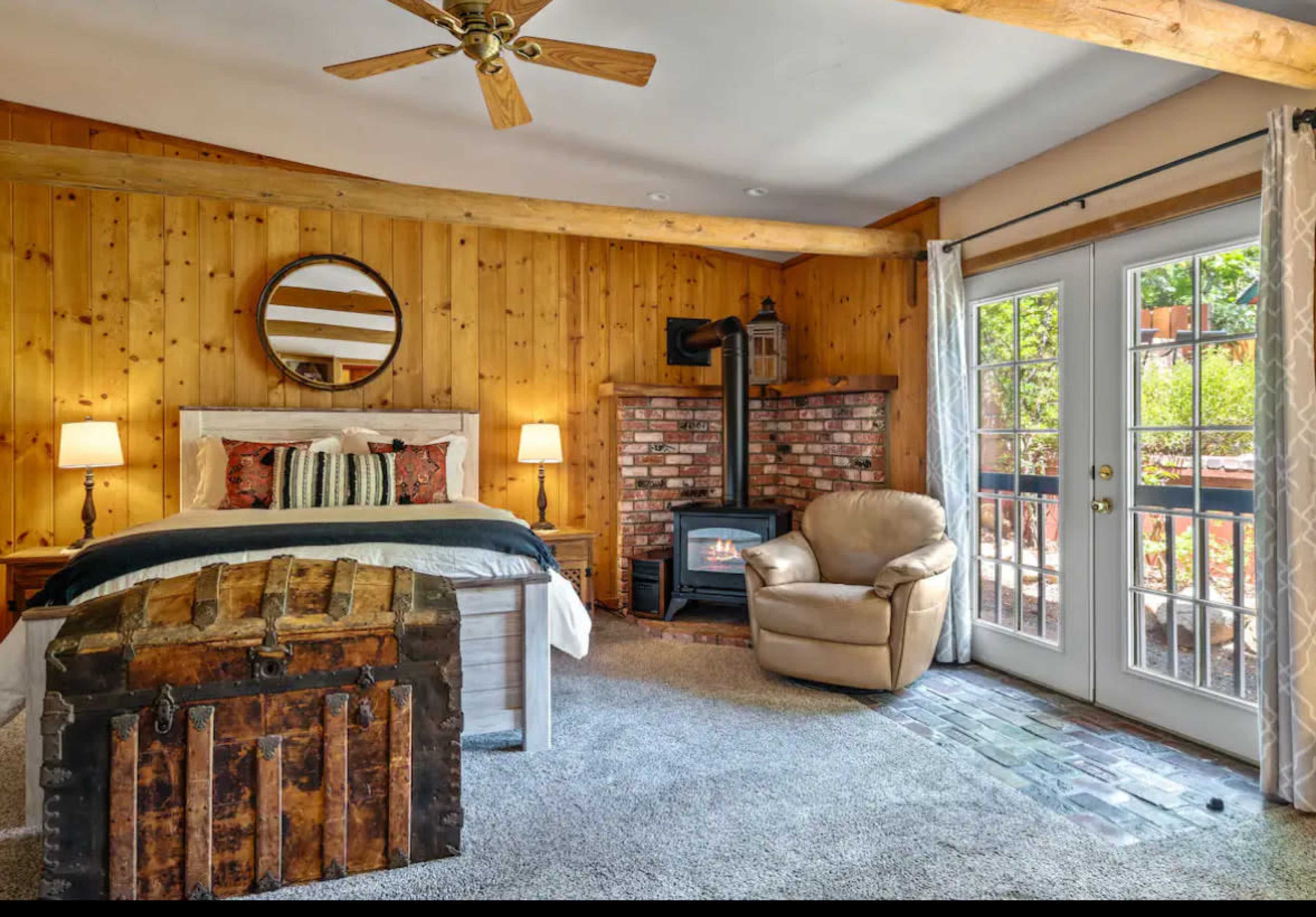 The image shows a cozy bedroom with wooden walls, a bed with decorative pillows, a brick fireplace, a round mirror, and a comfortable chair near a set of French doors leading to an outdoor area.