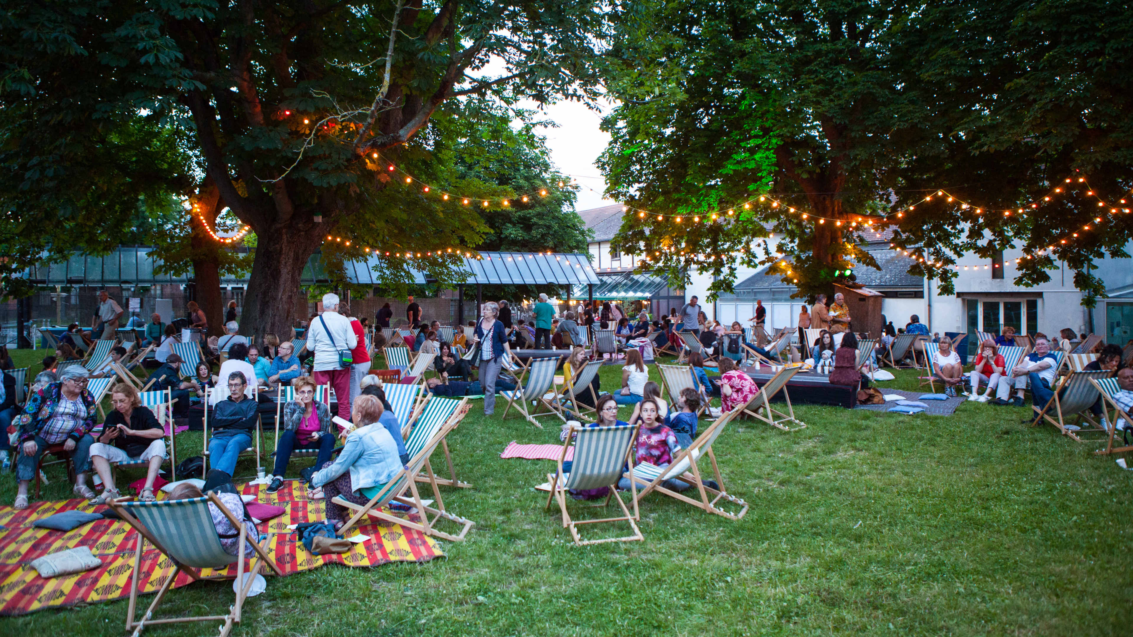 A large group of people gathers on a grassy area filled with deck chairs and blankets, enjoying an outdoor event illuminated by string lights.