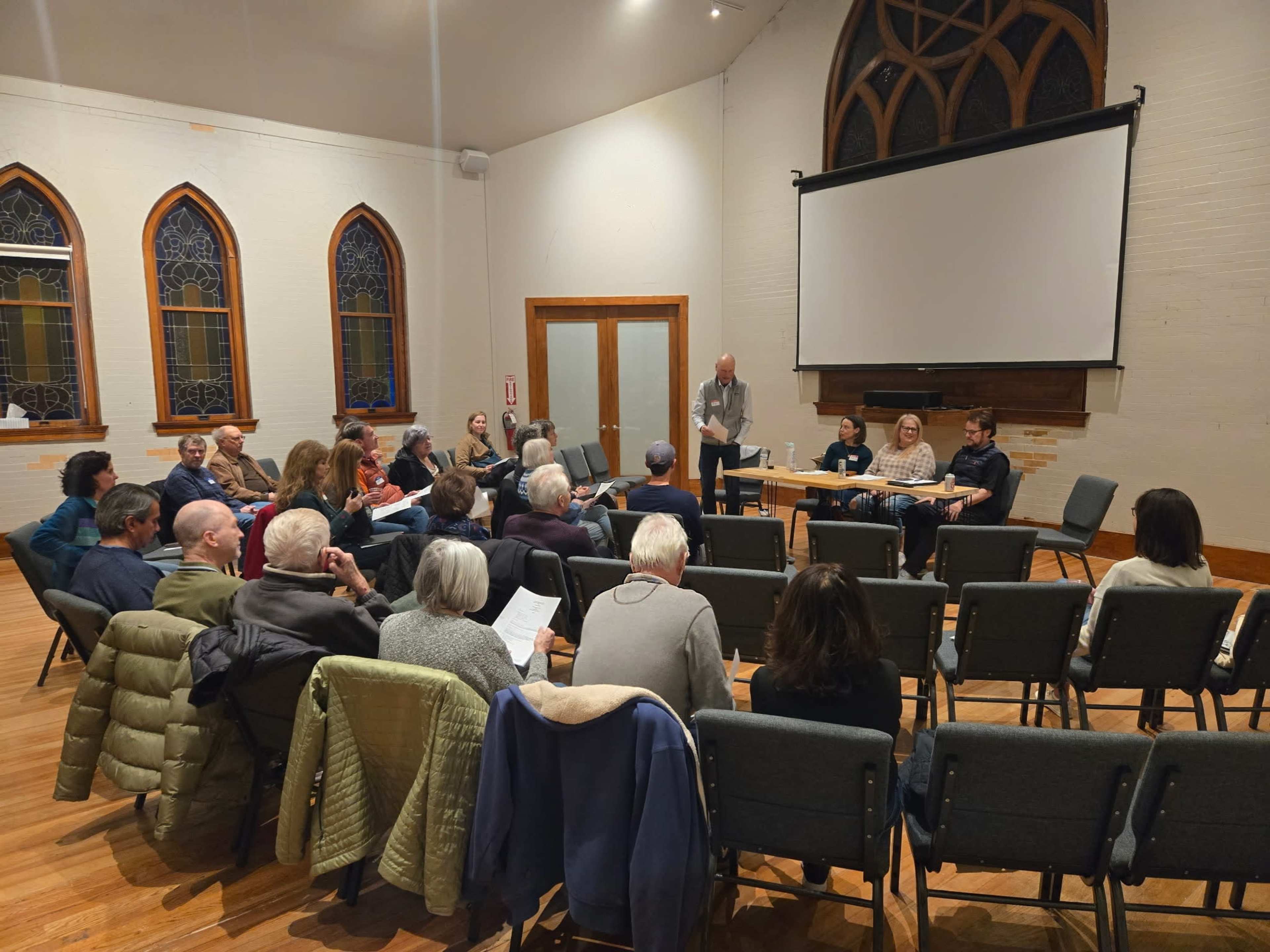 A group of people gathers in a hall with arched windows for a meeting, seated in rows facing a speaker at a table.