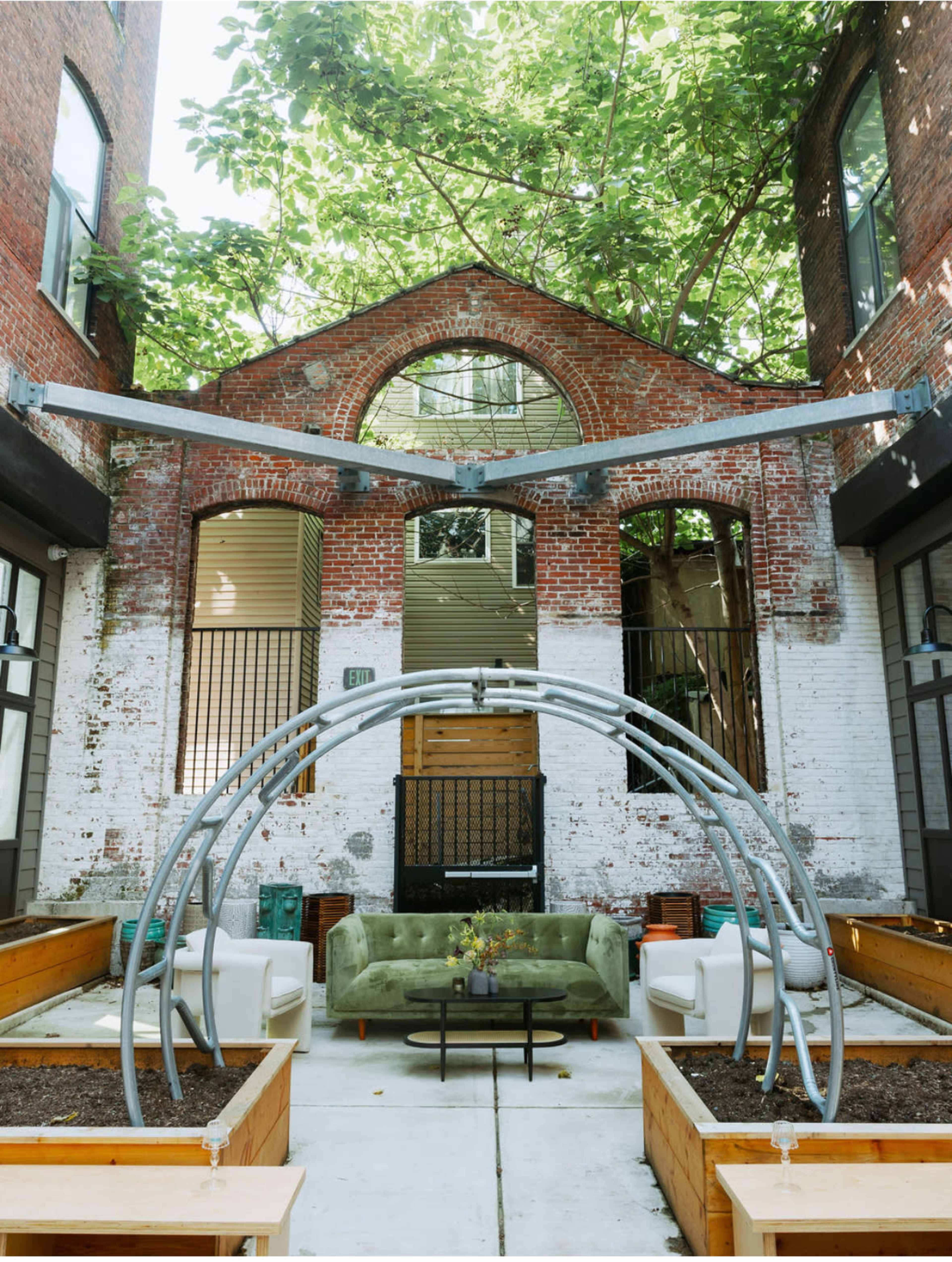 The image features a courtyard with exposed brick walls, a circular metal structure overhead, a green sofa, and white chairs surrounded by planters.