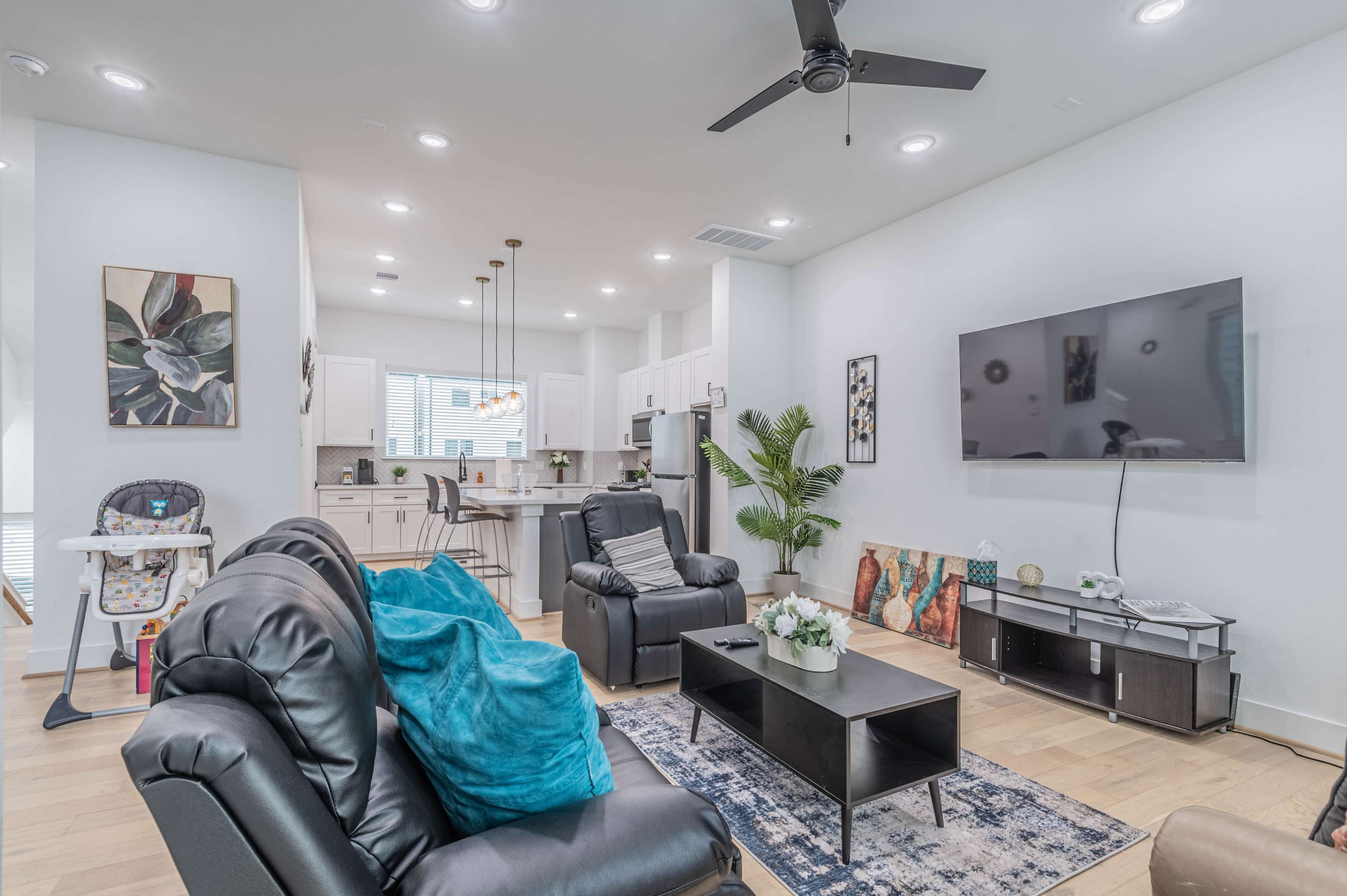 A modern living room with black recliners, a television mounted on the wall, a coffee table, and a kitchen area visible in the background.