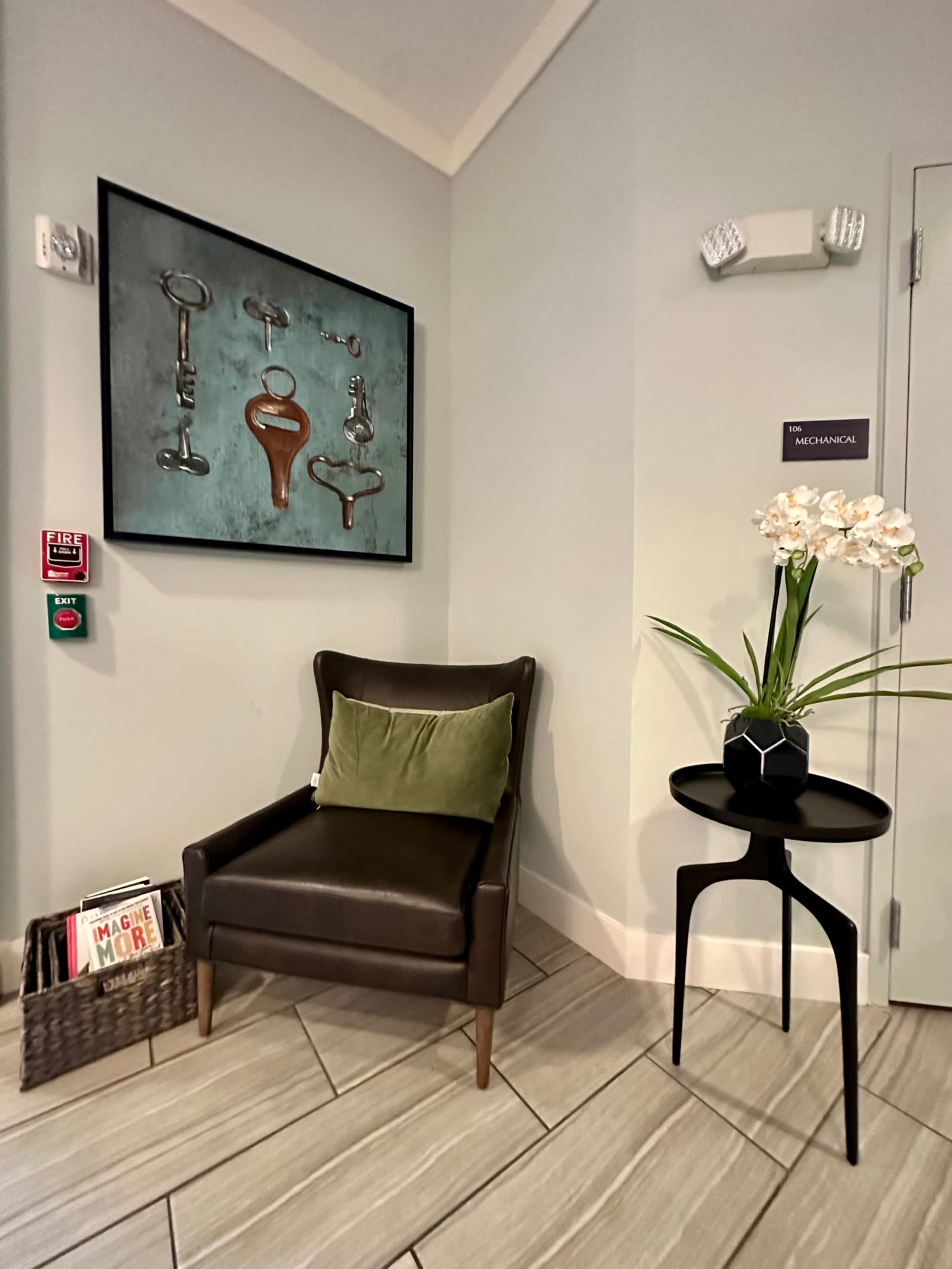 A corner of a room features a dark accent chair, a small black table with a plant, and artwork on the wall depicting various tools.