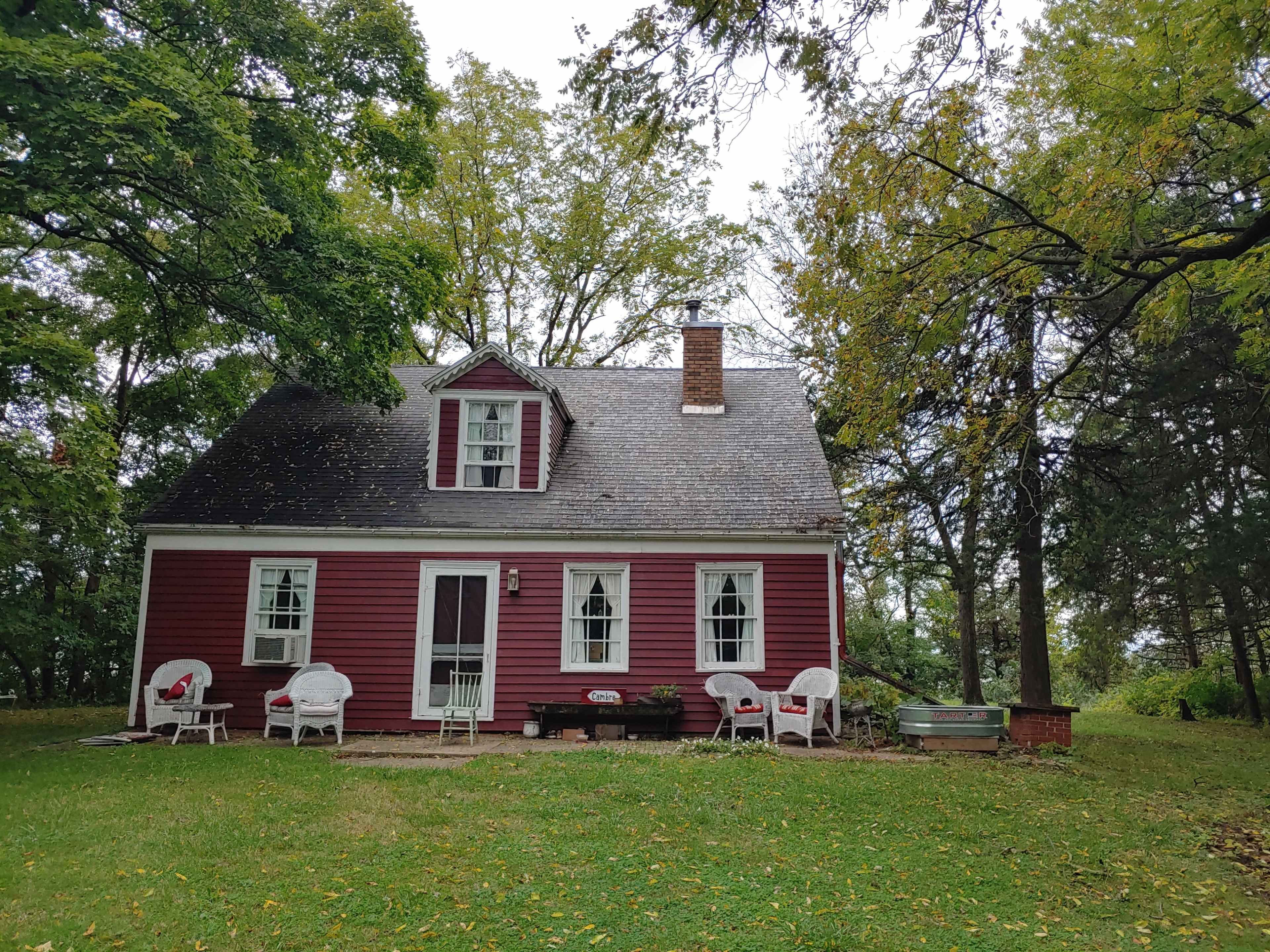 A red clapboard house with white trim sits surrounded by trees and lawn chairs on a grassy yard.