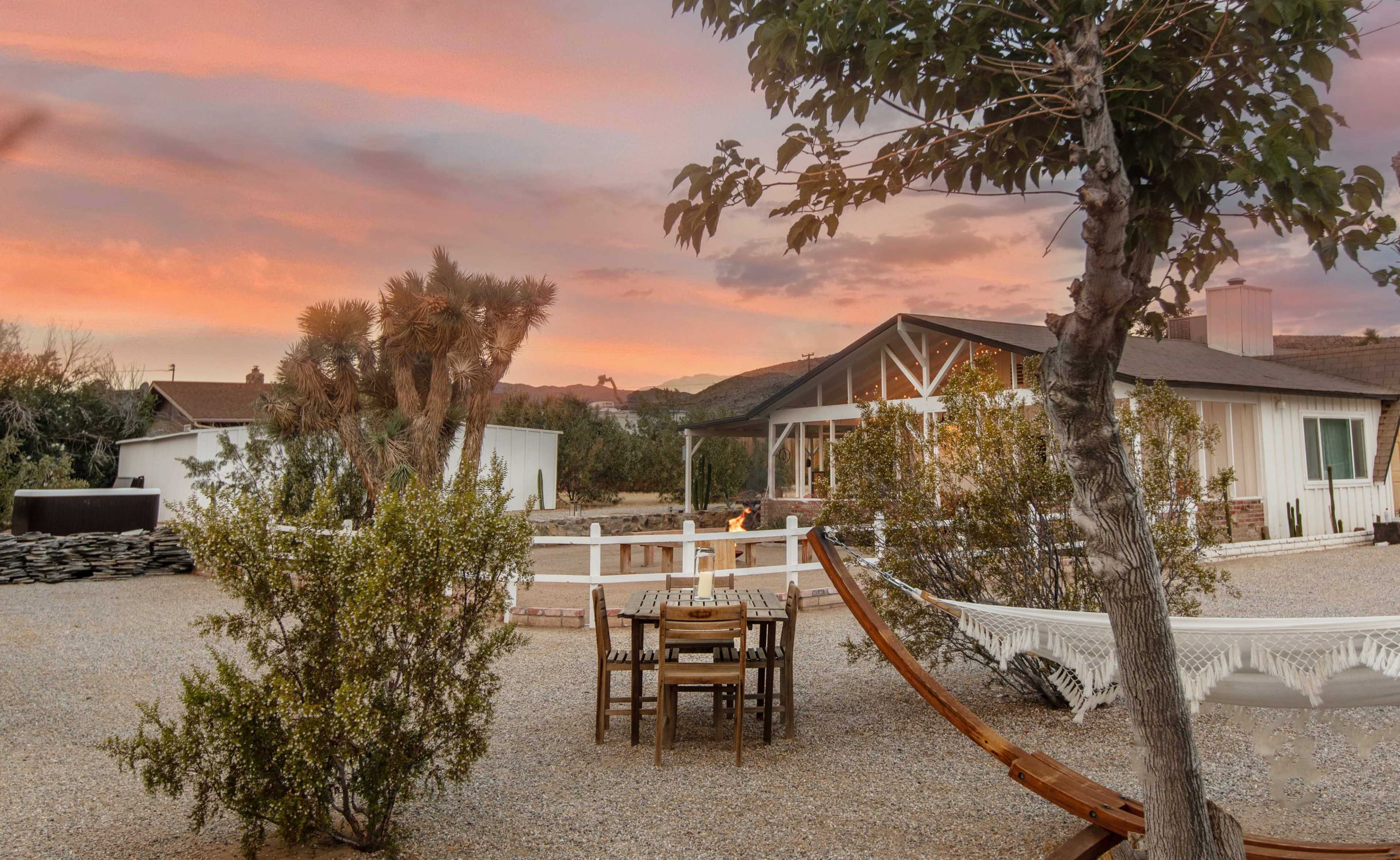 A wooden table and chairs sit beneath a tree in a gravel yard, with a hammock nearby and a house in the background against a colorful sunset sky.