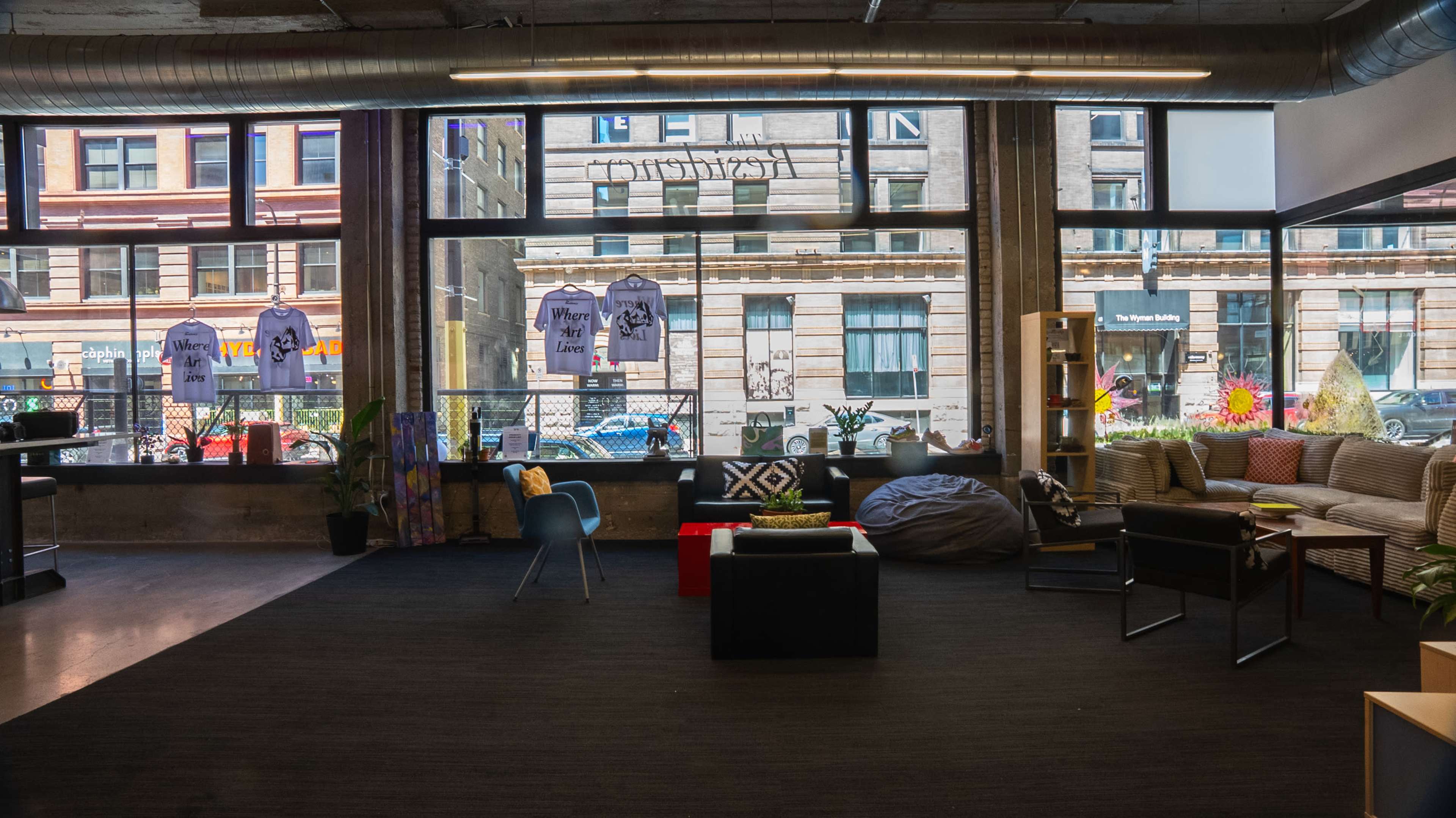 The image shows a cozy interior space with seating, plants, and artwork, featuring large windows that open to a street view with hanging shirts displayed outside.