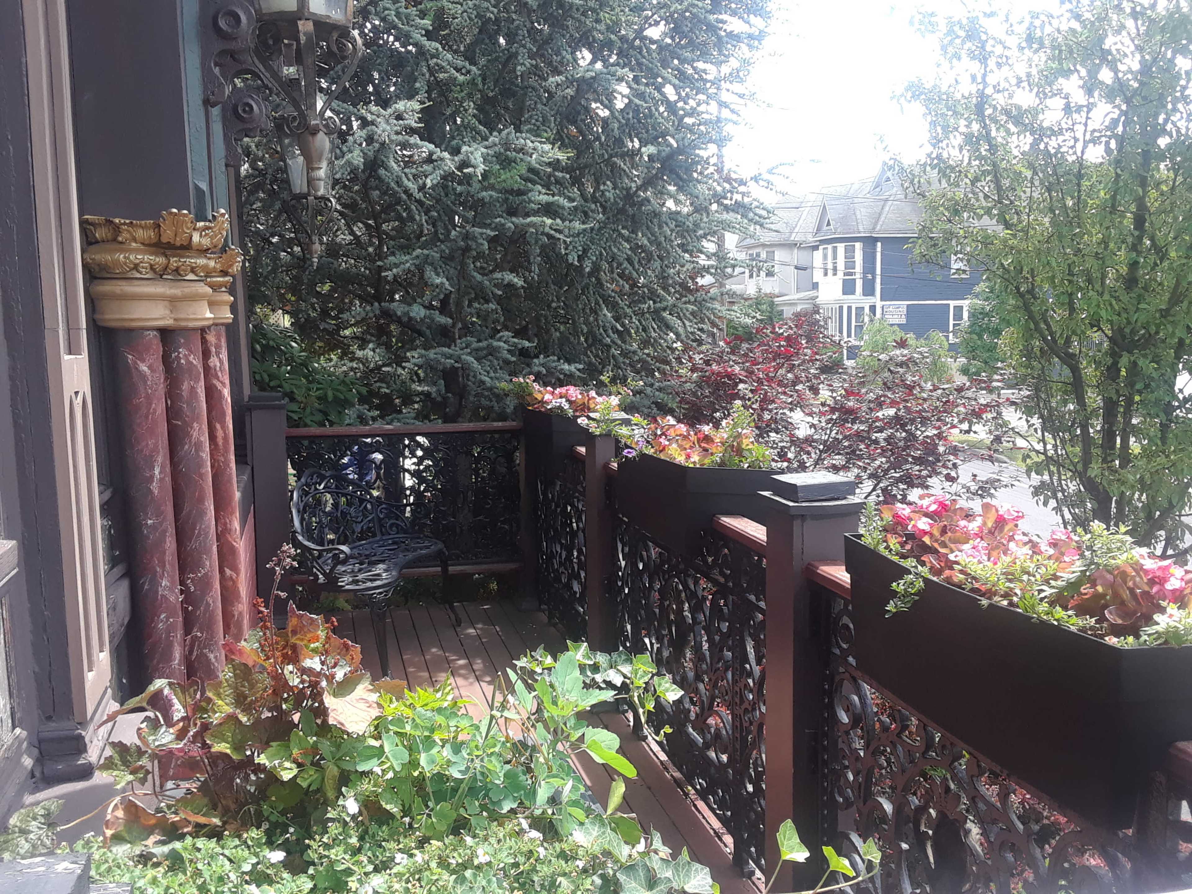 A balcony with decorative railings and planters filled with greenery overlooks a garden and nearby houses.