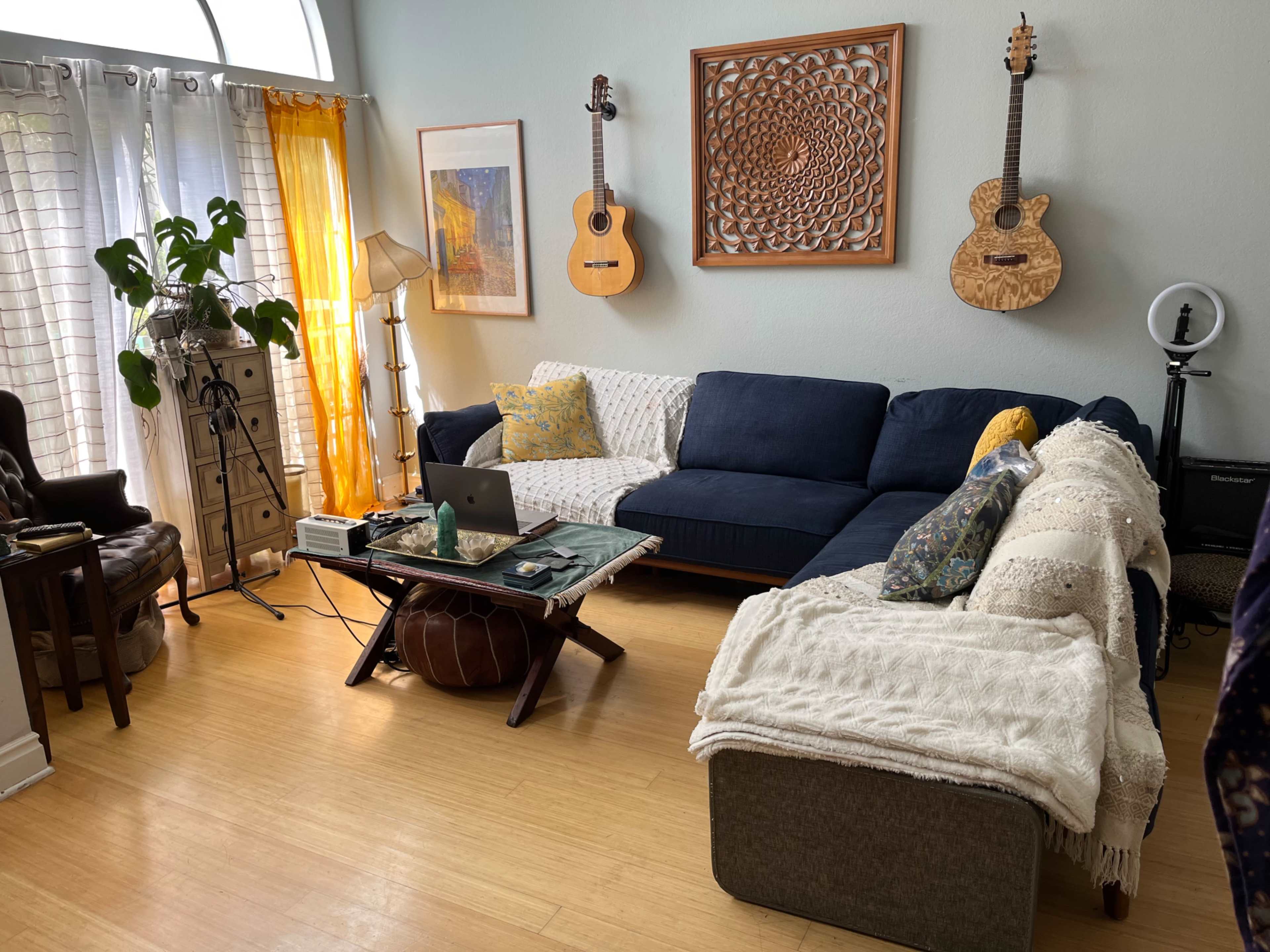 A cozy living room features a blue sectional sofa, a wooden coffee table with a laptop, two guitars on the wall, and a potted plant by the window.