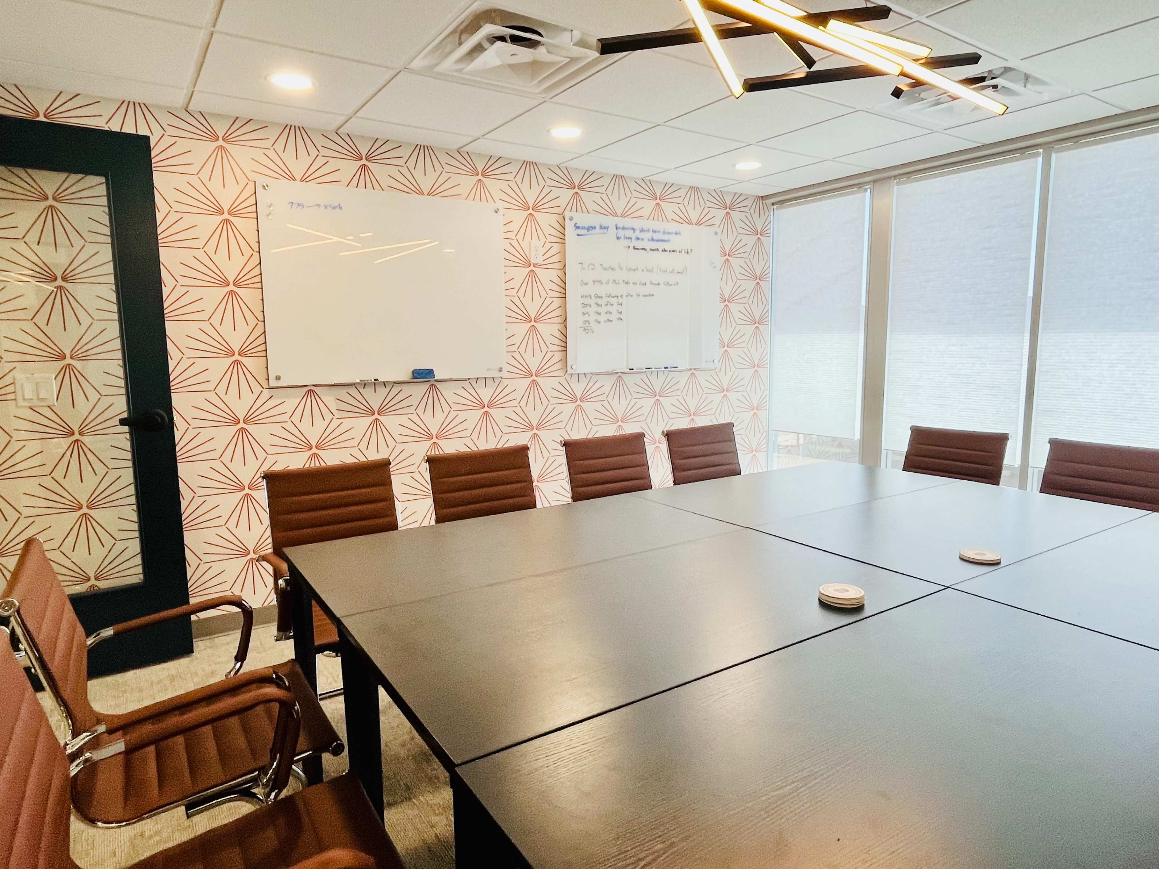 A modern conference room features a large table surrounded by brown leather chairs, with whiteboards on the walls and natural light streaming in through large windows.
