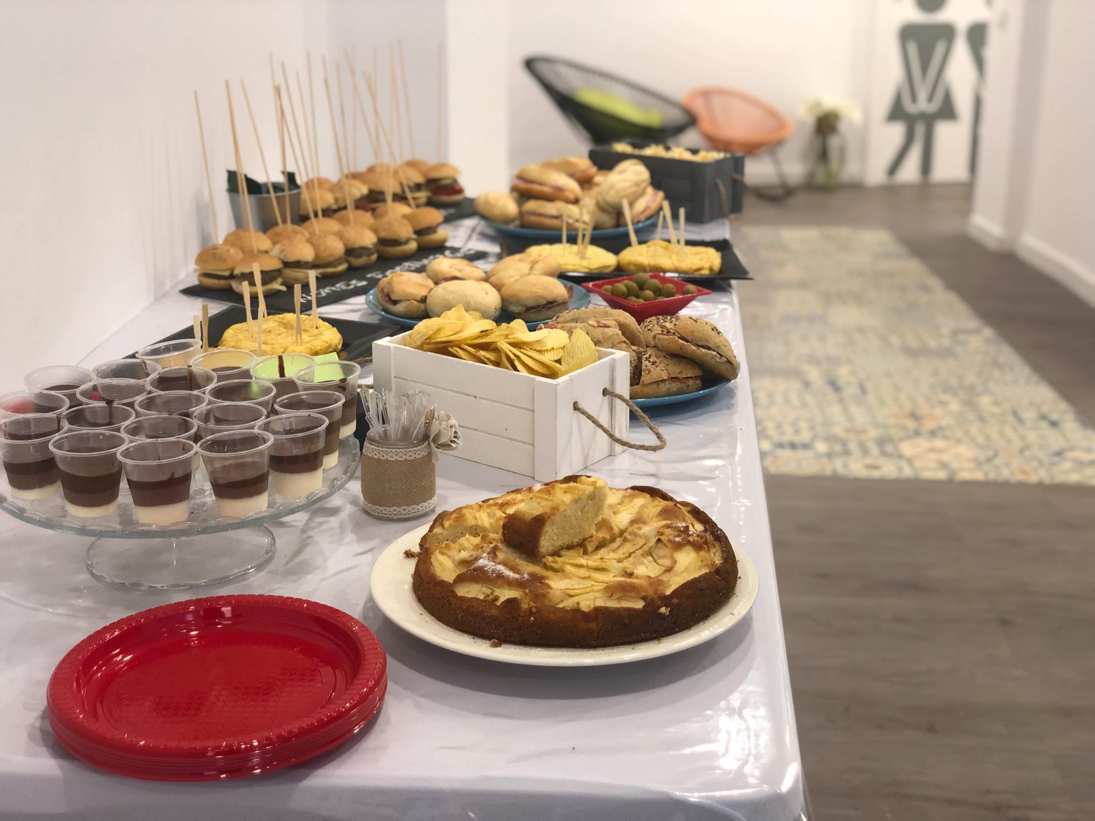 A table is set with various food items, including burgers, snacks, desserts, and drinks in clear cups.