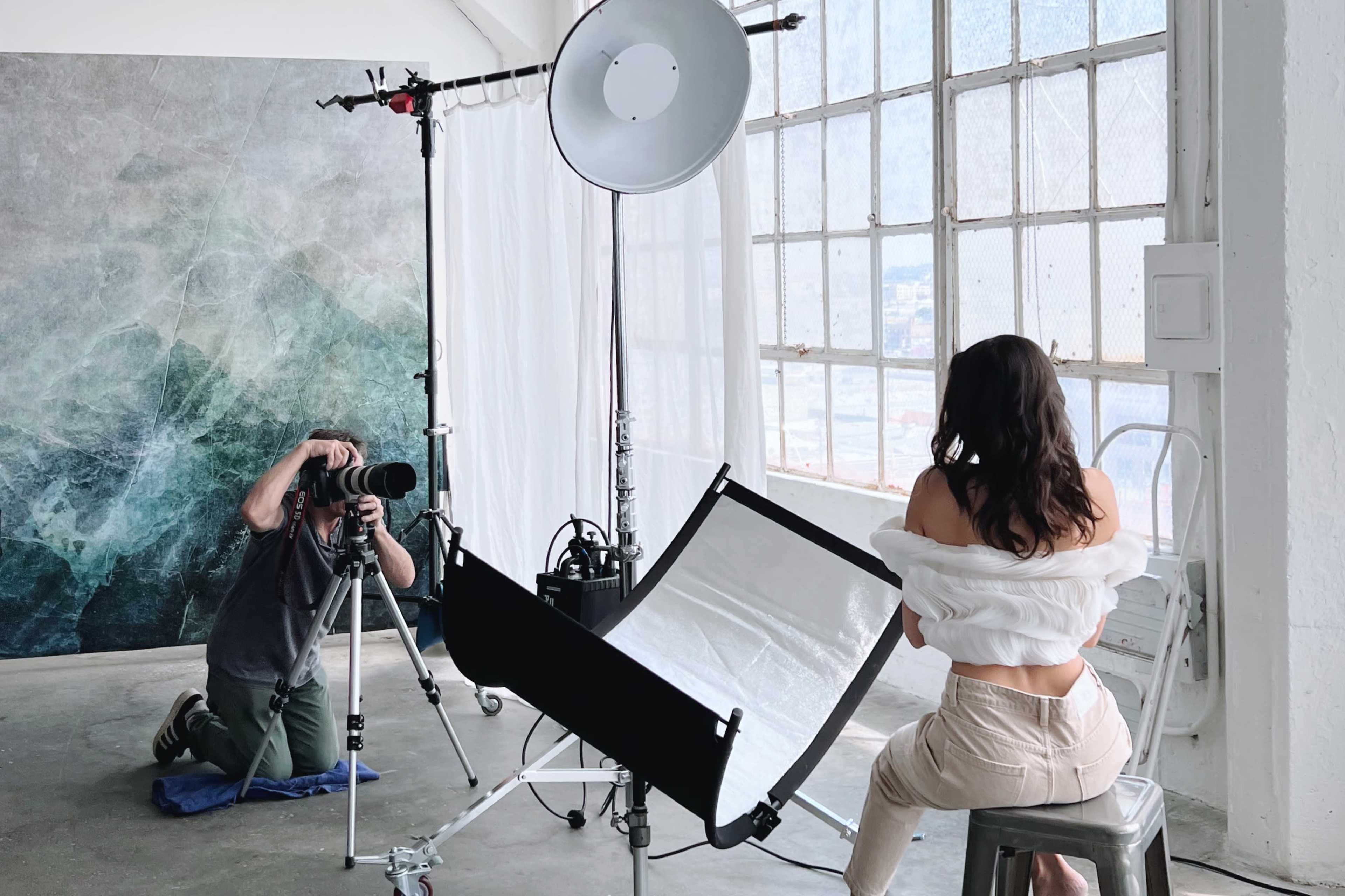 A photographer captures images of a model in a studio setting with large windows and a backdrop.