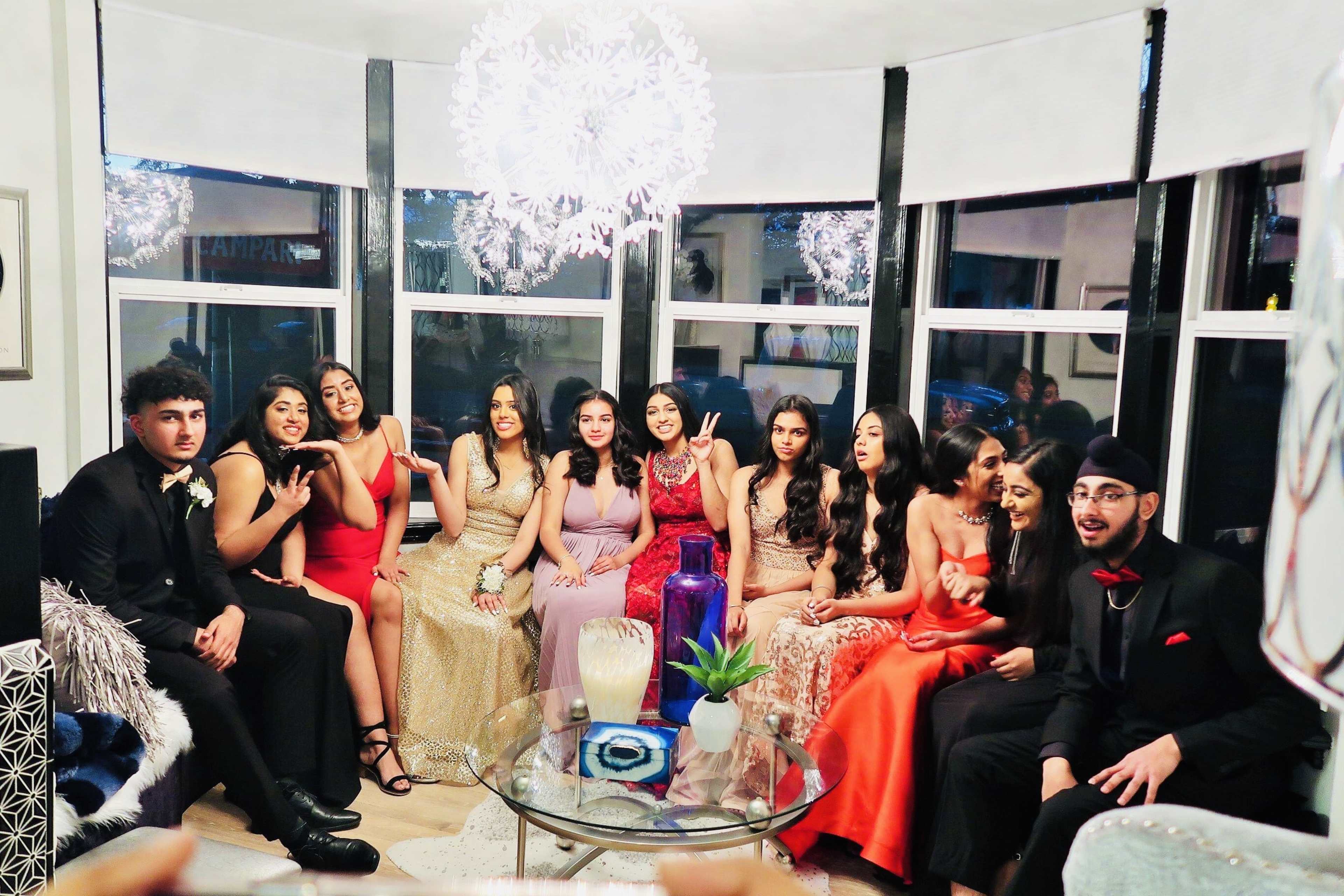 A group of thirteen young adults in formal attire poses together in a decorated living room with large windows and modern lighting.