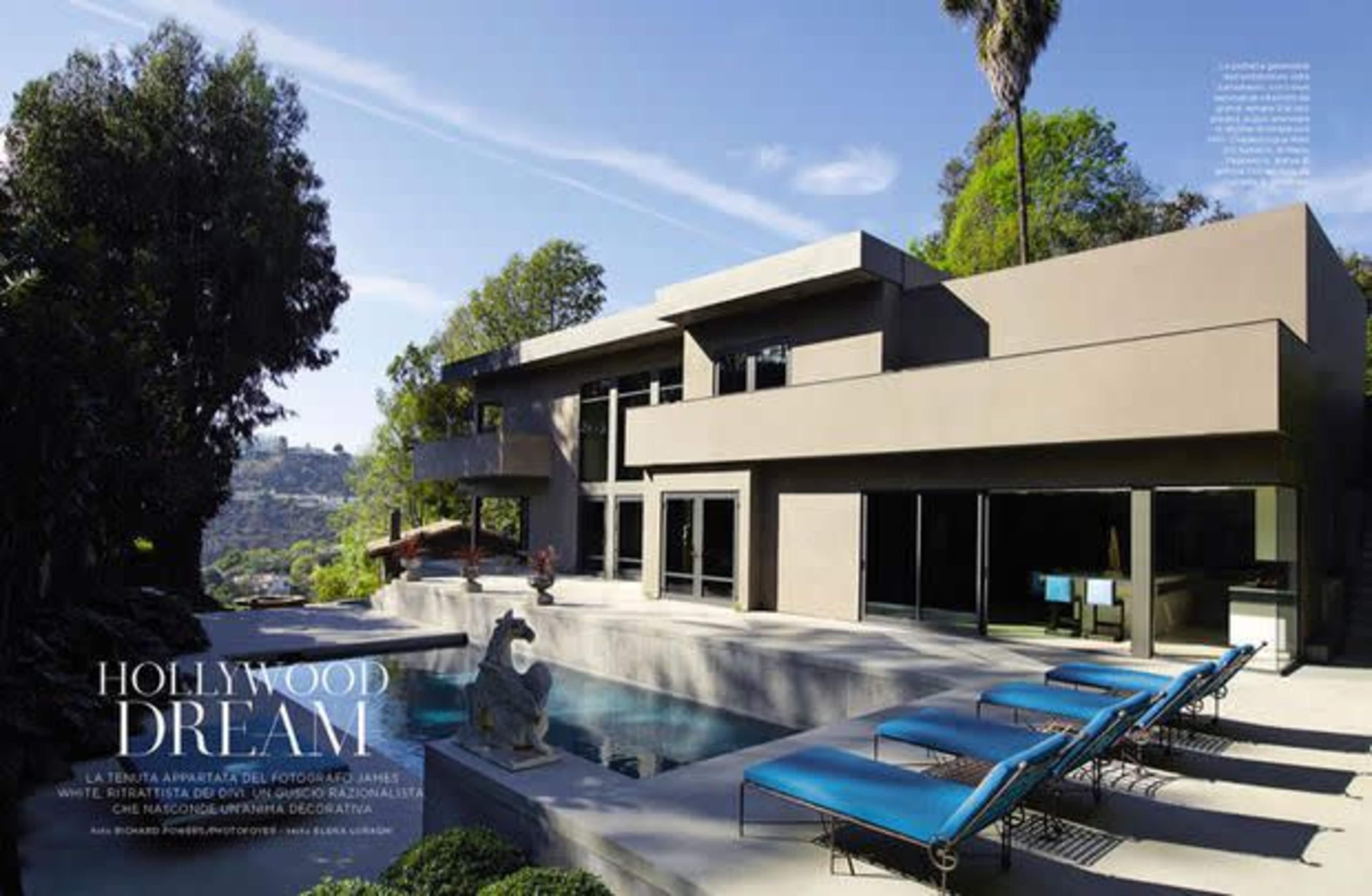 A modern house with large windows overlooks a swimming pool and landscaped garden in a hillside setting.