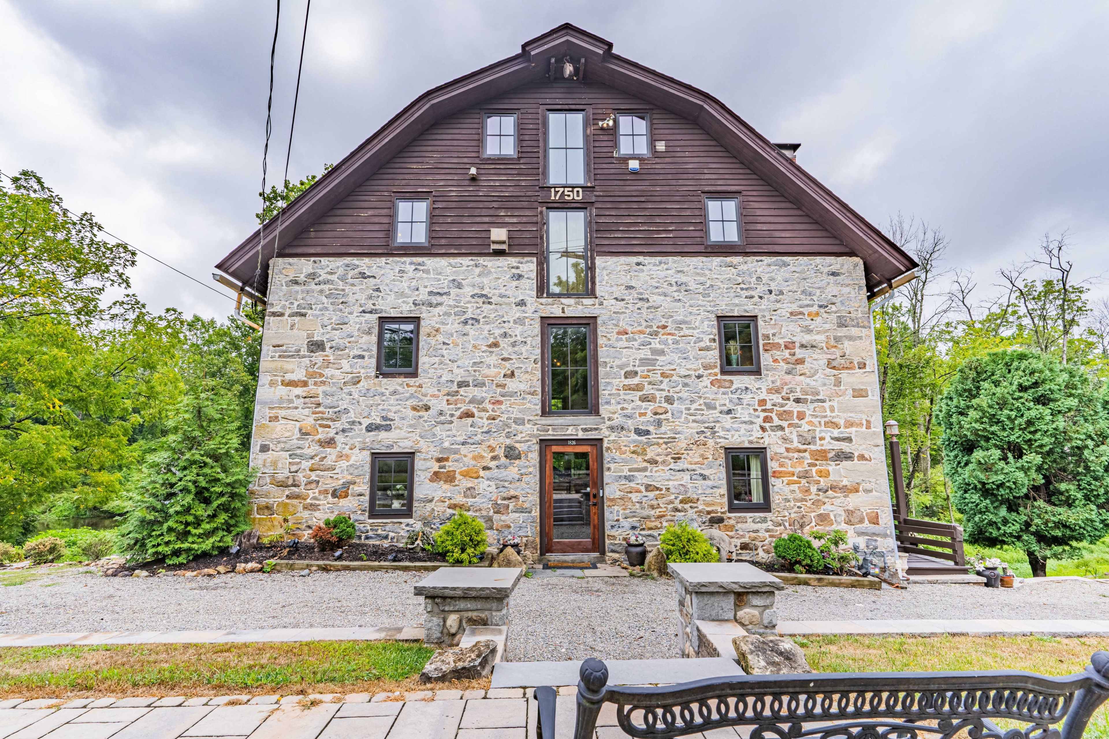 A three-story stone building with a triangular roof has a wooden upper section, set in a landscaped area with a gravel pathway.