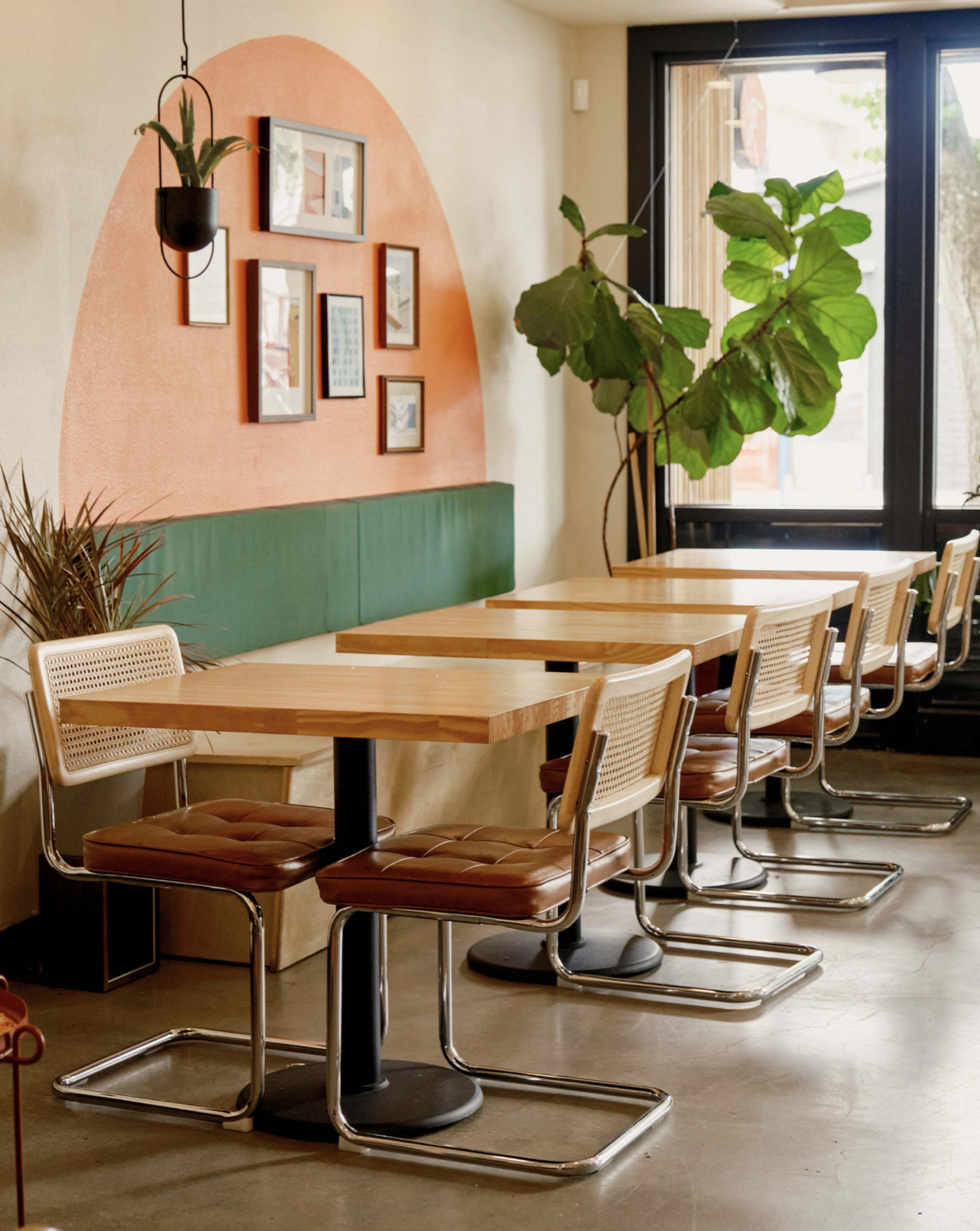 The image shows a modern café interior with several wooden tables and chairs arranged along a wall featuring an arched mural and framed pictures.