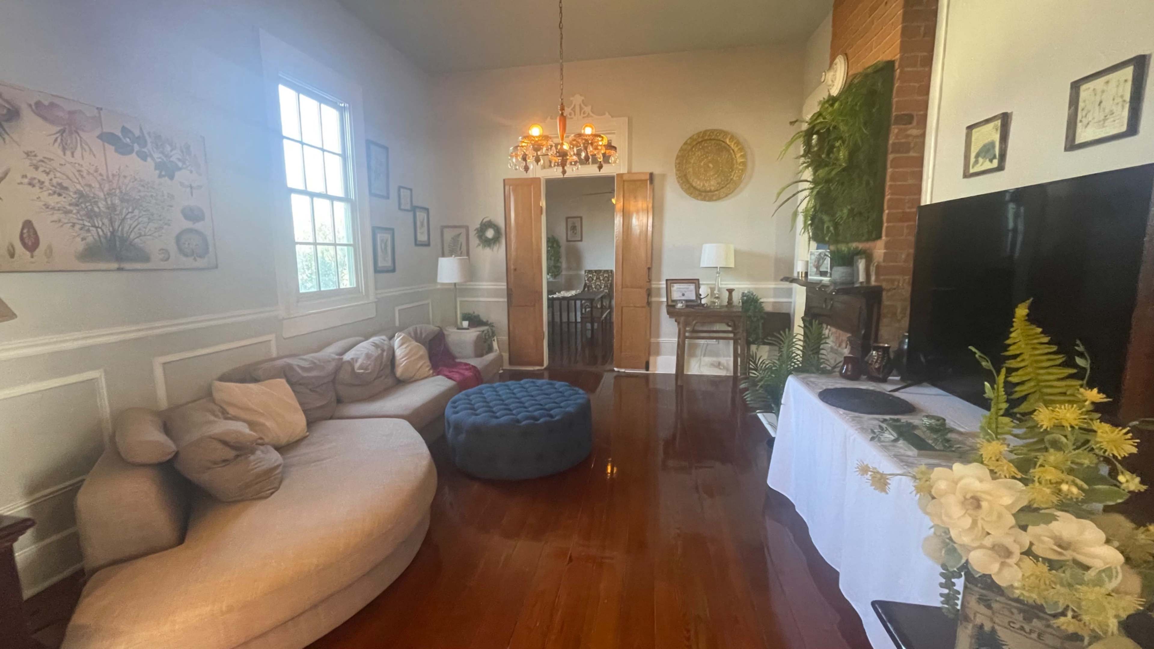 The image shows a cozy living room featuring a light-colored sectional sofa, a round ottoman, hardwood floors, and various decorative elements including plants and artwork on the walls.