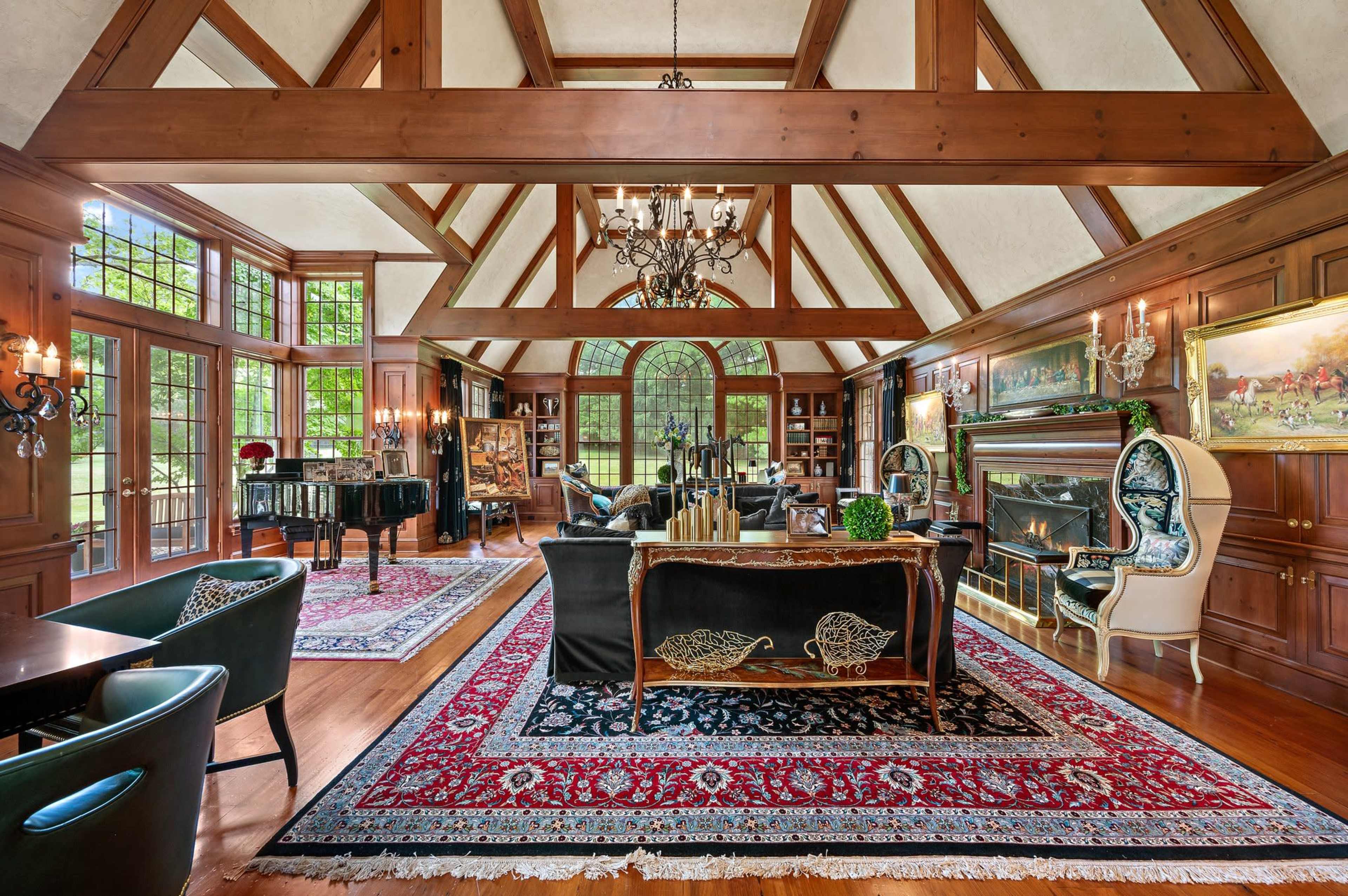 The image shows an elegantly designed living room with wooden beams, a grand piano, a fireplace, and large windows allowing natural light to fill the space.